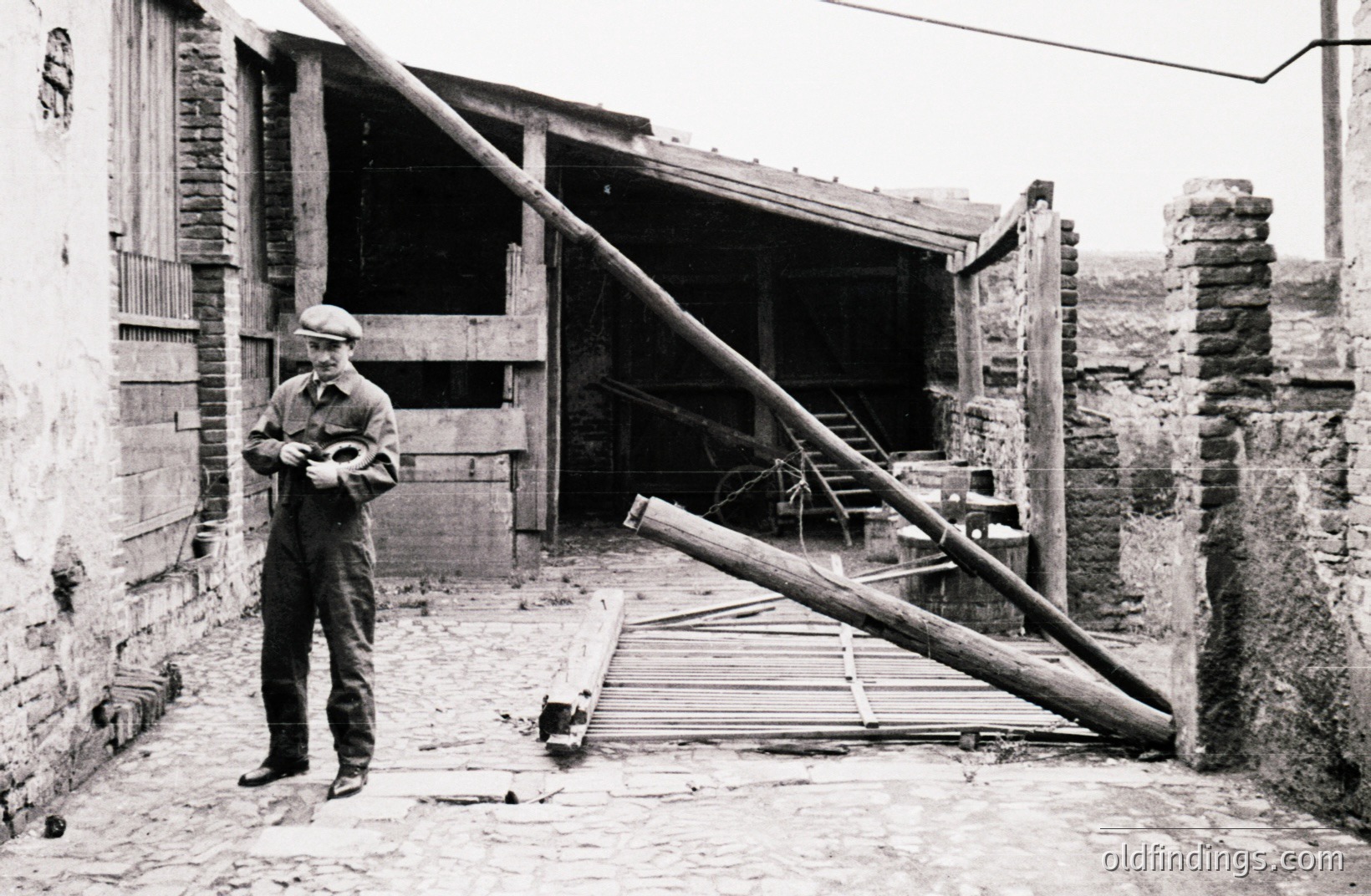 Man in work overalls and cap stands in a brick courtyard, examining a small object in his hands. Weathered brick walls and a partially visible wooden structure define the space. A large timber log rests against a gate. Likely a rural or industrial setting, possibly 1940s-1960s.