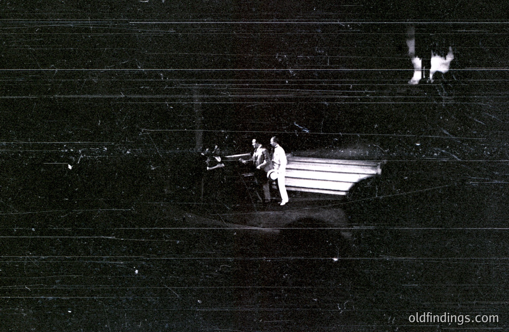 A monochrome shot depicts two men examining a large, angular architectural model. The model, likely a building facade, is rendered in stark relief. The setting seems to be an interior space, indicated by the dark background. Likely a design studio or construction site.