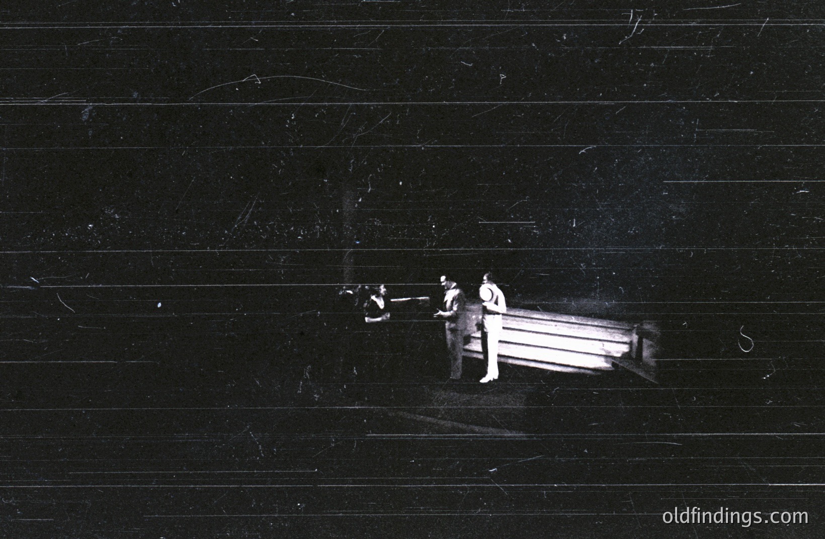 Three figures stand near a rectangular structure, possibly a platform or stage, partially obscured in darkness. The scene appears staged or performance-related. Grainy monochrome imagery suggests mid-20th century photography. Likely a theatrical or experimental art context. Minimal visible details offer location clues.