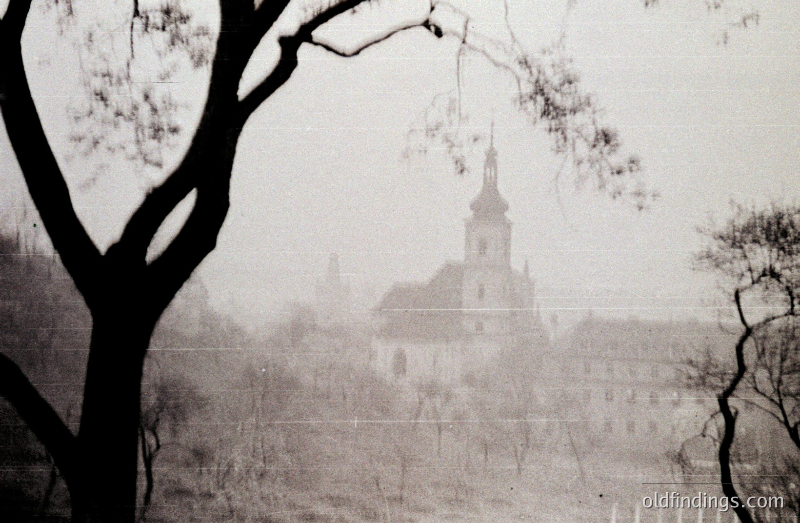 A view of a historic townscape, likely European, shrouded in mist. A church spire dominates the scene, visible above rooftops. Silhouetted tree branches frame the composition. Appears to be a vintage photographic print, displaying age-related grain and surface texture. Possible 1950s-1970s era.