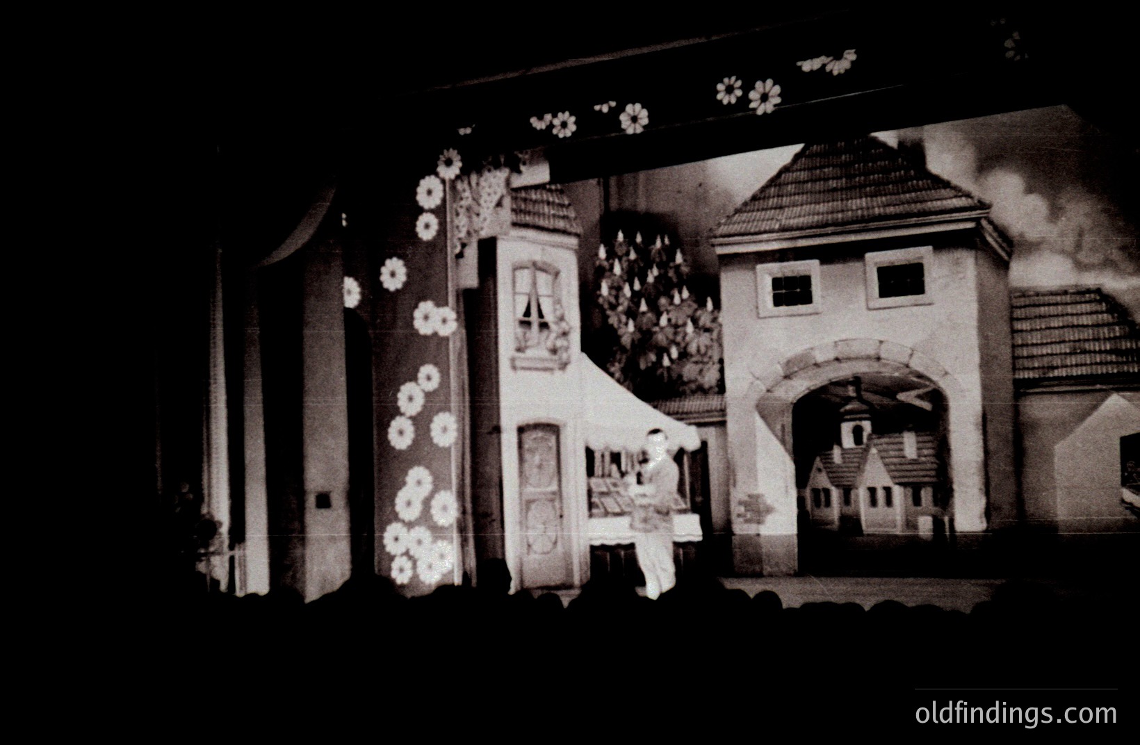 A monochromatic staged scene depicting a fairytale or folk village. A figure in period attire stands before an ornate gateway, flanked by buildings with detailed facades and pointed roofs. Decorative floral motifs adorn the structures. Likely a theatrical backdrop, c. 1930s-1950s.