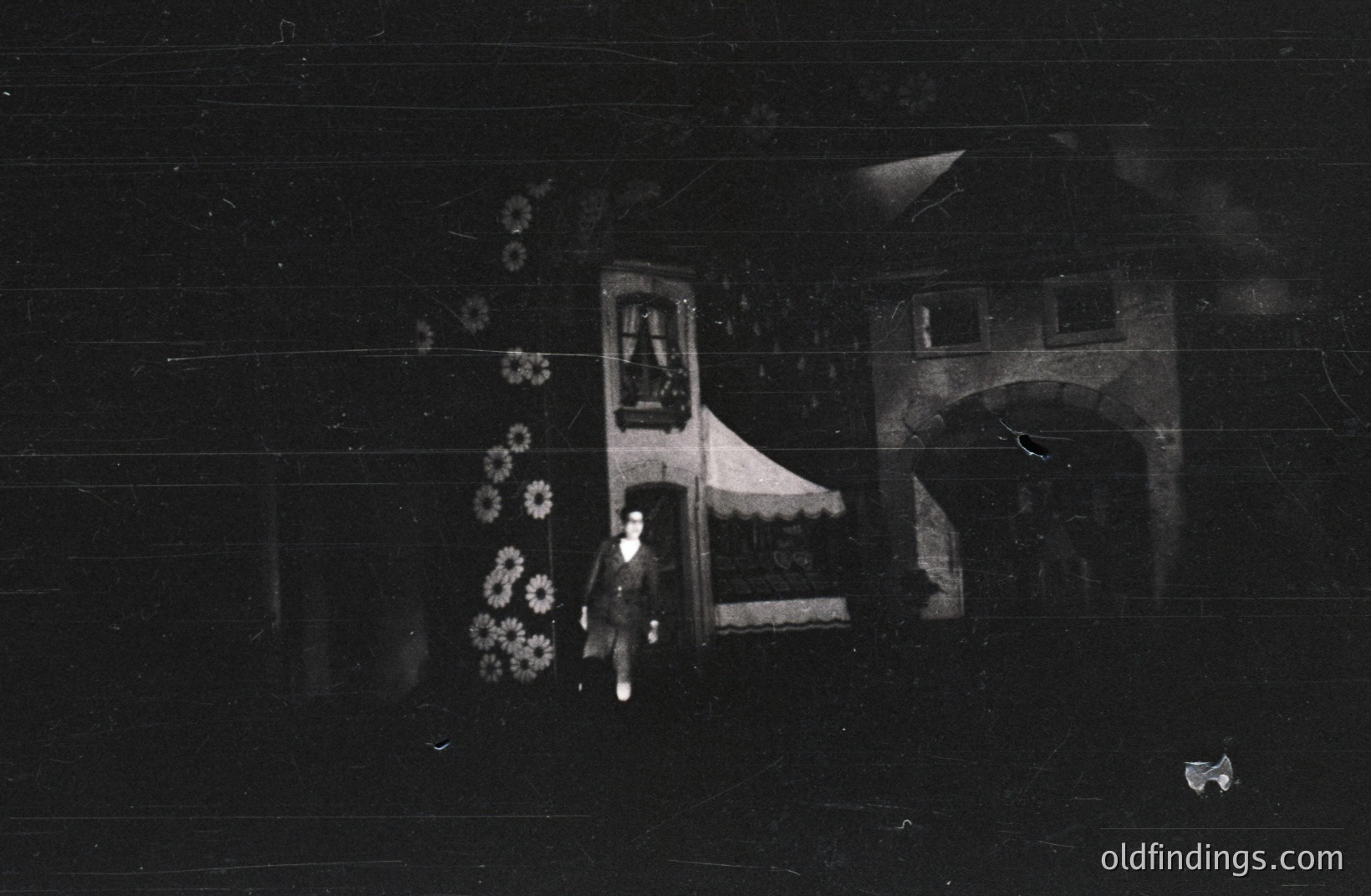 A monochrome image shows a person standing near a building featuring a detailed balcony with ornate railings and a partially visible arched entrance. The building’s architecture suggests a European style, possibly early 20th century. Dense floral motifs decorate the facade. Overall mood is serene, with aged film grain adding texture.
