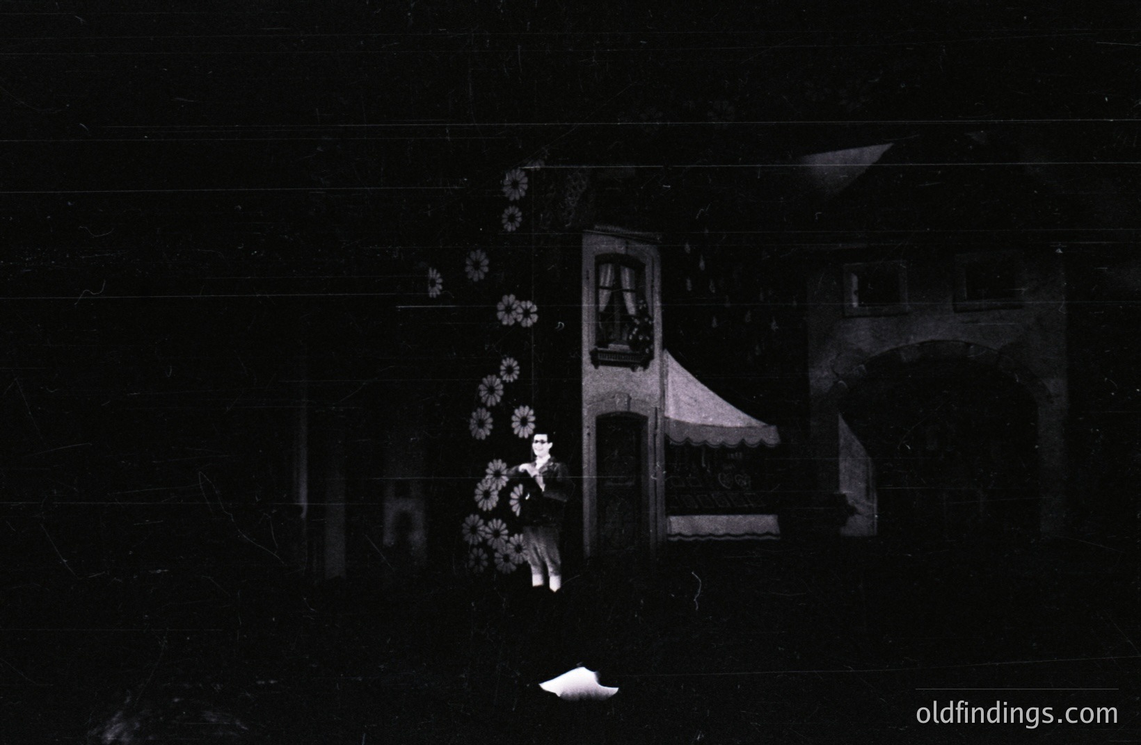 A young woman stands before a building facade, likely a theatrical set. The structure includes a balcony, arched entryway, and striped awning. The scene features minimal lighting, suggesting a stage or backdrop. Likely mid-20th century, possibly a film or play still.