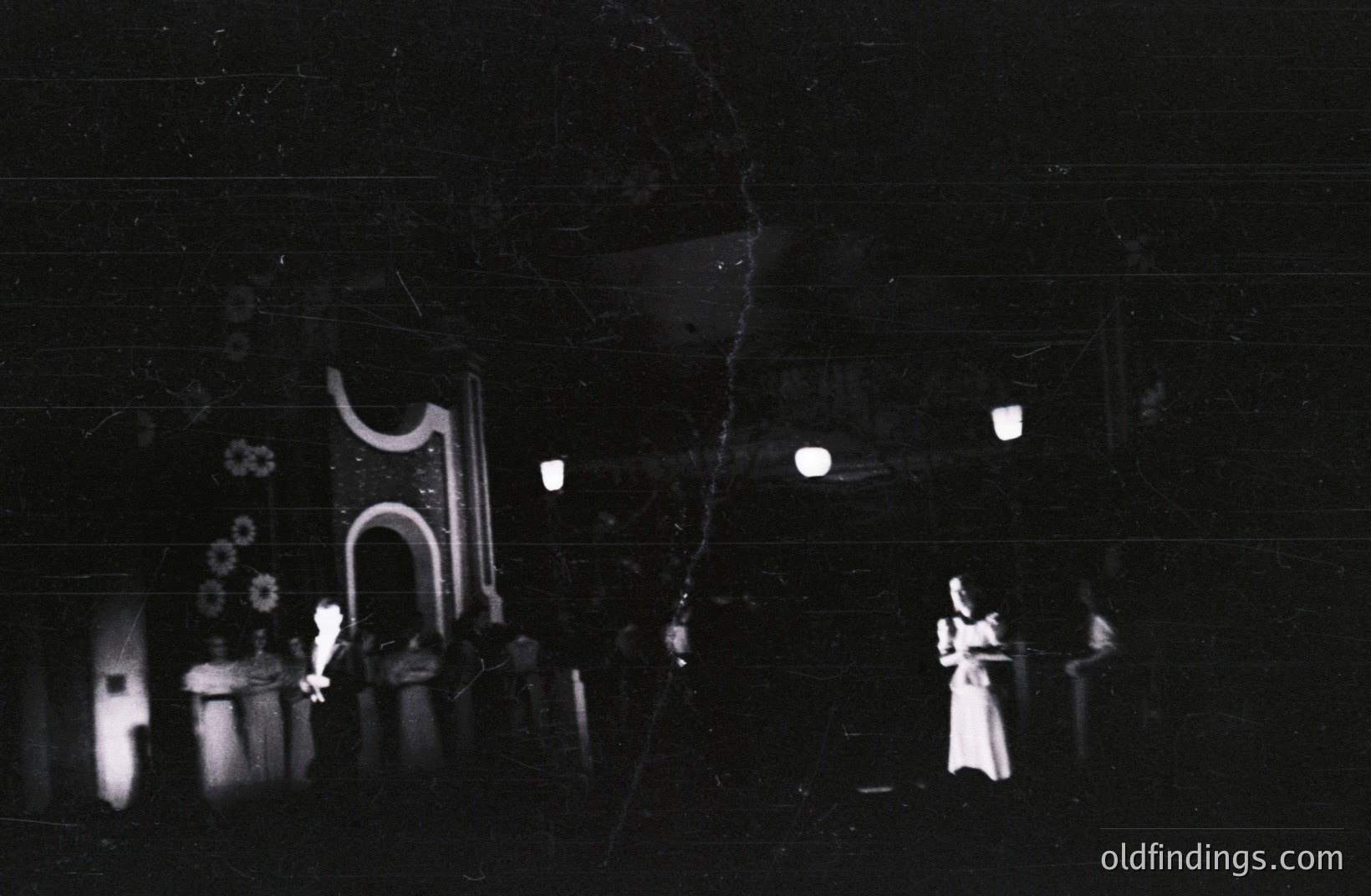 Eerie scene of a theatrical performance, possibly outdoors. A figure in white stands center, surrounded by draped fabric & stylized architectural elements. Lanterns provide dim lighting, enhancing the dramatic effect. Appears to be late 1960s-70s.