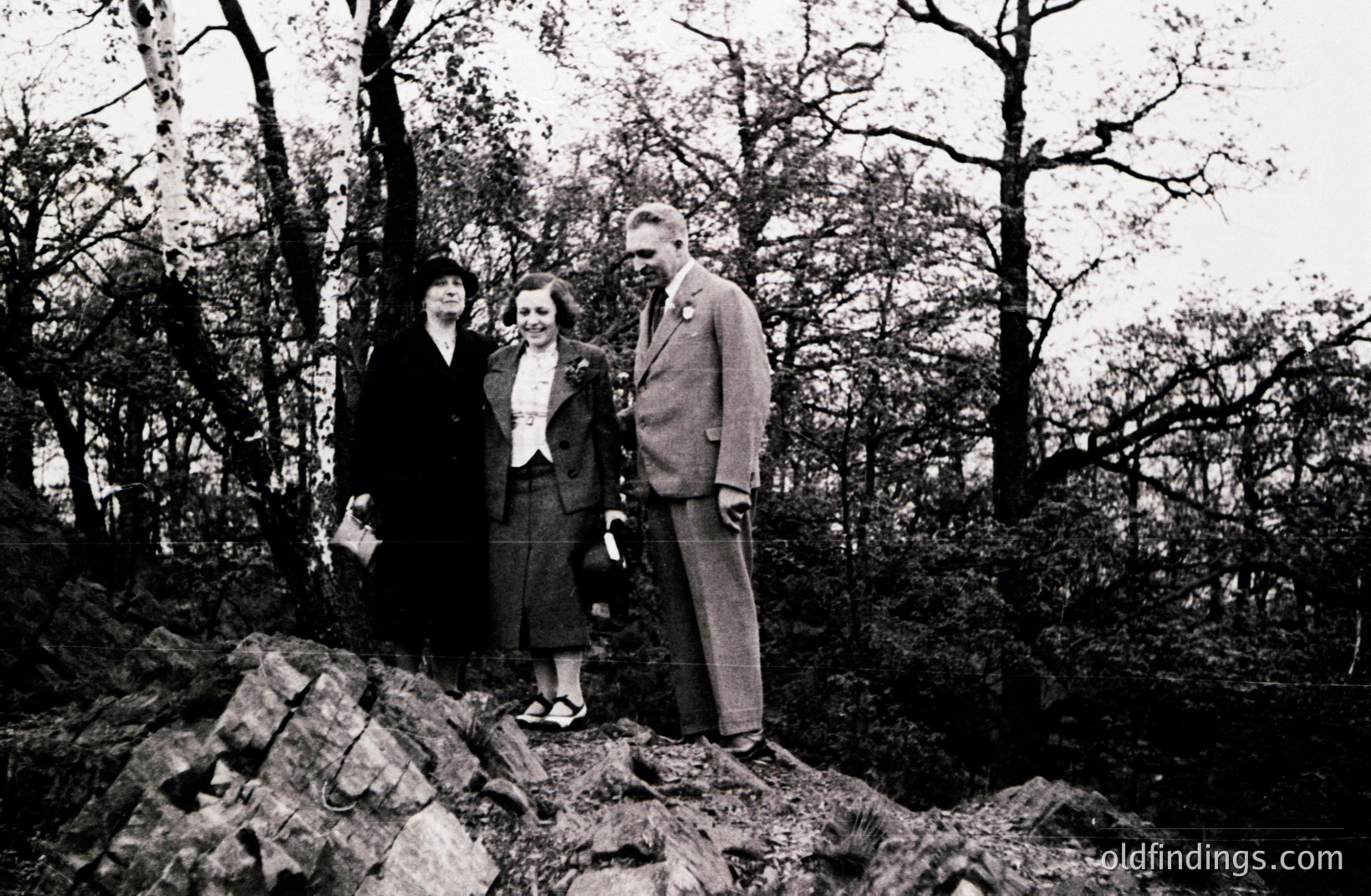 Three figures pose on a rocky outcrop amidst dense trees. A woman in a dark suit and hat stands to the left; a younger woman in a tailored suit and floral embellishment stands center, and a man in a suit and tie to the right. Likely a formal portrait from the 1940s.