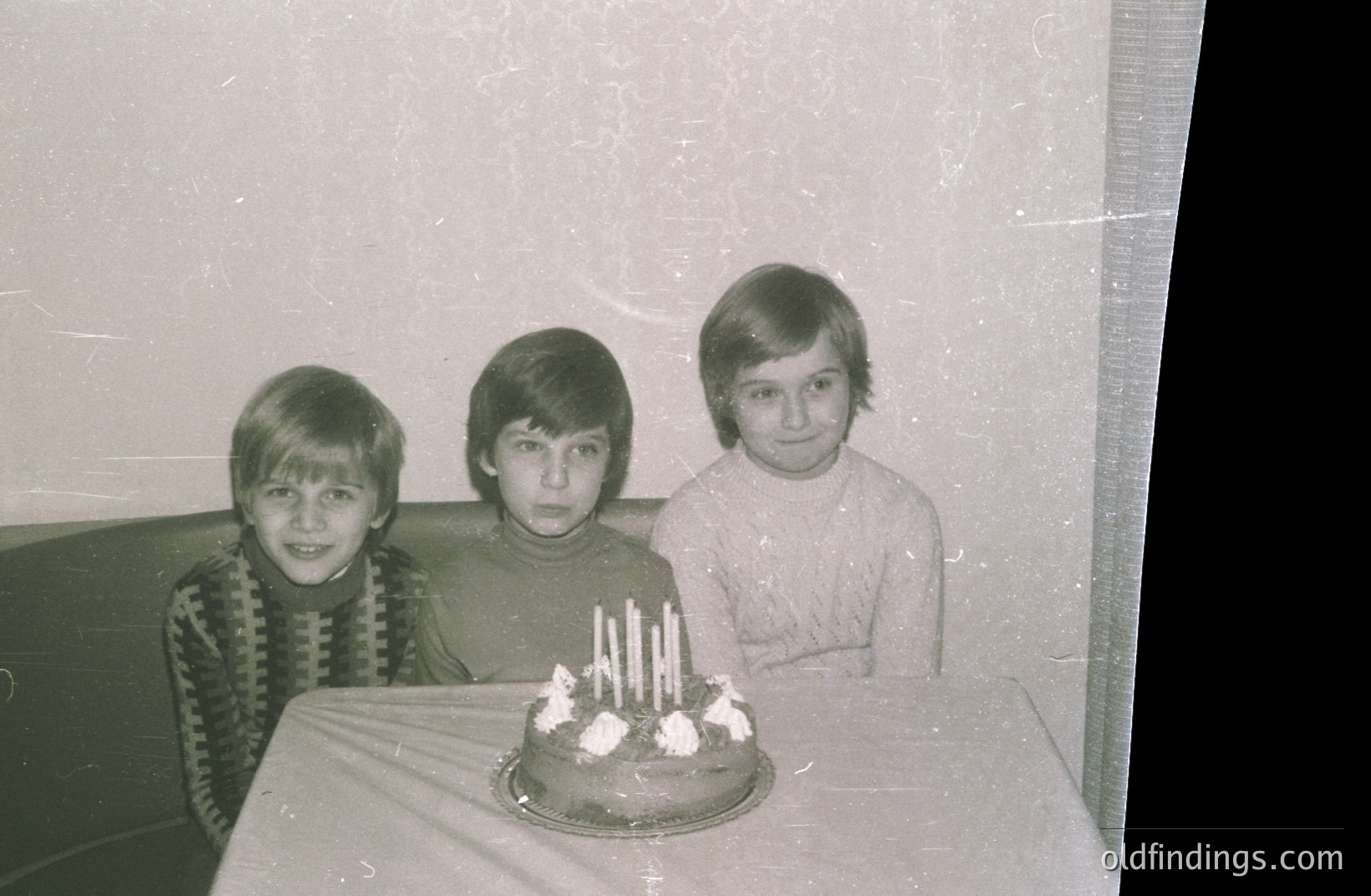 Three boys, likely siblings, sit behind a cake with lit candles. The image shows a mid-century style interior. Clothing suggests a 1970s aesthetic, with turtlenecks & patterned sweaters. The photograph has aged, showing signs of wear and vintage paper quality.
