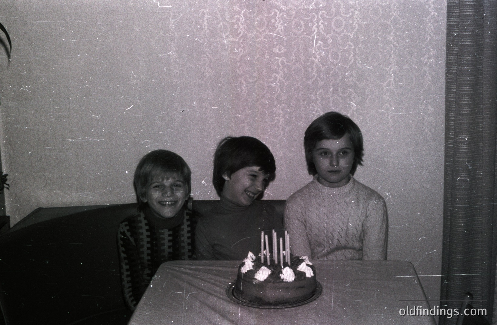 Three young boys, likely siblings, pose with a chocolate cake adorned with lit candles in a domestic setting. Visible details include textured wallpaper, a draped table, and vintage clothing. The grainy monochrome suggests a 1970s time period. A charming, candid birthday moment.