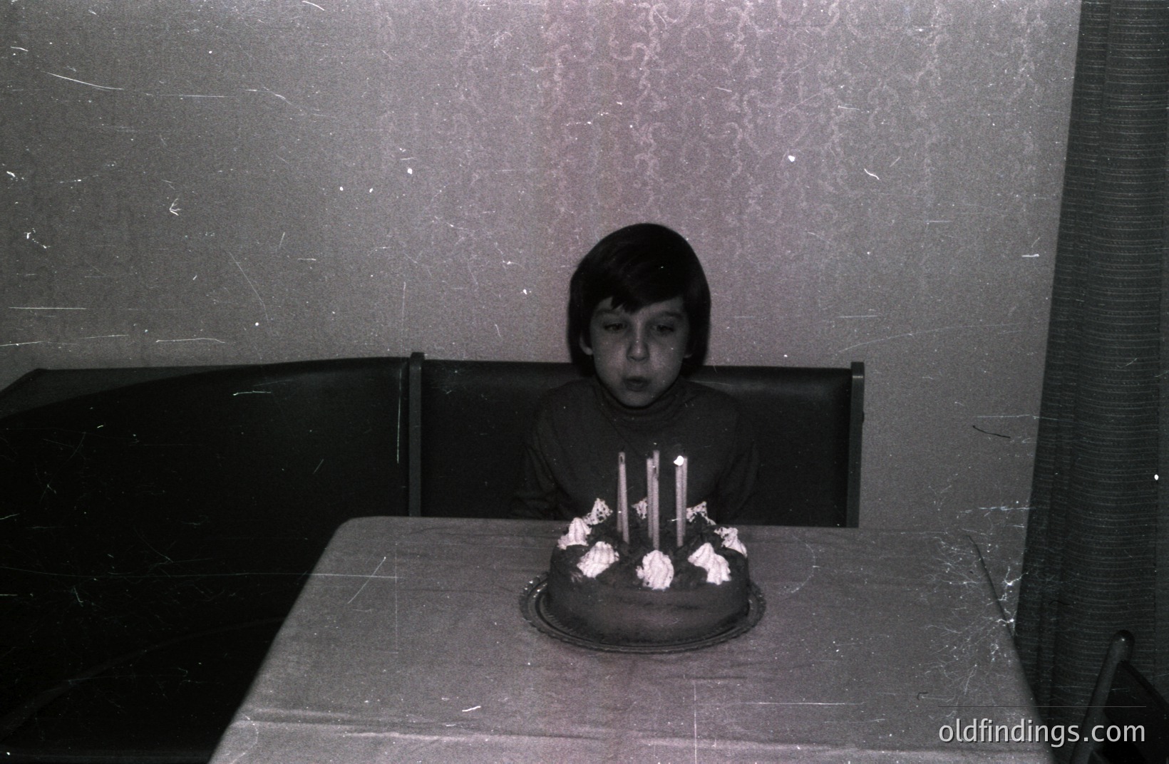 A young boy sits at a table covered with a tablecloth, focused on a birthday cake with seven lit candles. The room features textured wallpaper and vintage furniture, indicative of a likely 1970s interior. Excellent for nostalgia projects or historical family archives. The photograph’s condition shows age-related signs.
