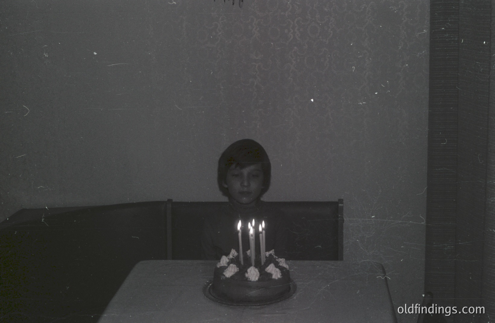 A young person sits behind a table covered with a tablecloth, facing a cake with seven candles. The room's wallpaper features a patterned design. Likely a personal snapshot, indicative of a mid-century domestic scene. The image's texture suggests vintage film stock.
