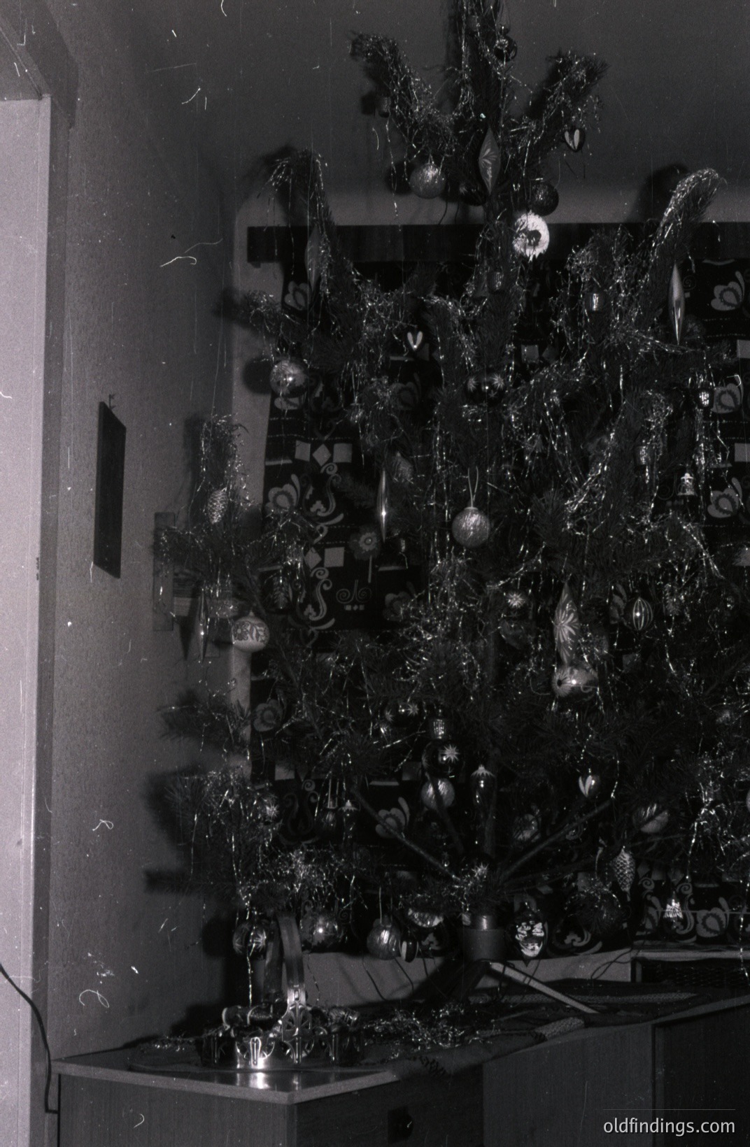 A festive, flocked artificial Christmas tree, heavily adorned with silver tinsel and ornaments, dominates this indoor scene. The tree stands on what appears to be a sideboard or cabinet. Likely from the 1960s, reflecting popular holiday décor styles. Appears to be a domestic interior.