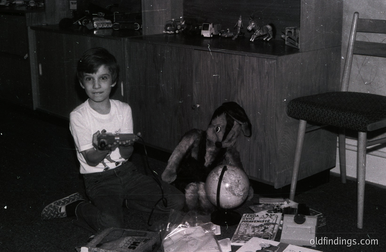 A young boy sits amidst toys and books, clutching a toy rifle. He's positioned in front of a dark wood cabinet displaying miniature vehicles. Likely a snapshot from the 1960s or 70s, showcasing childhood play. Nostalgic family archive material.