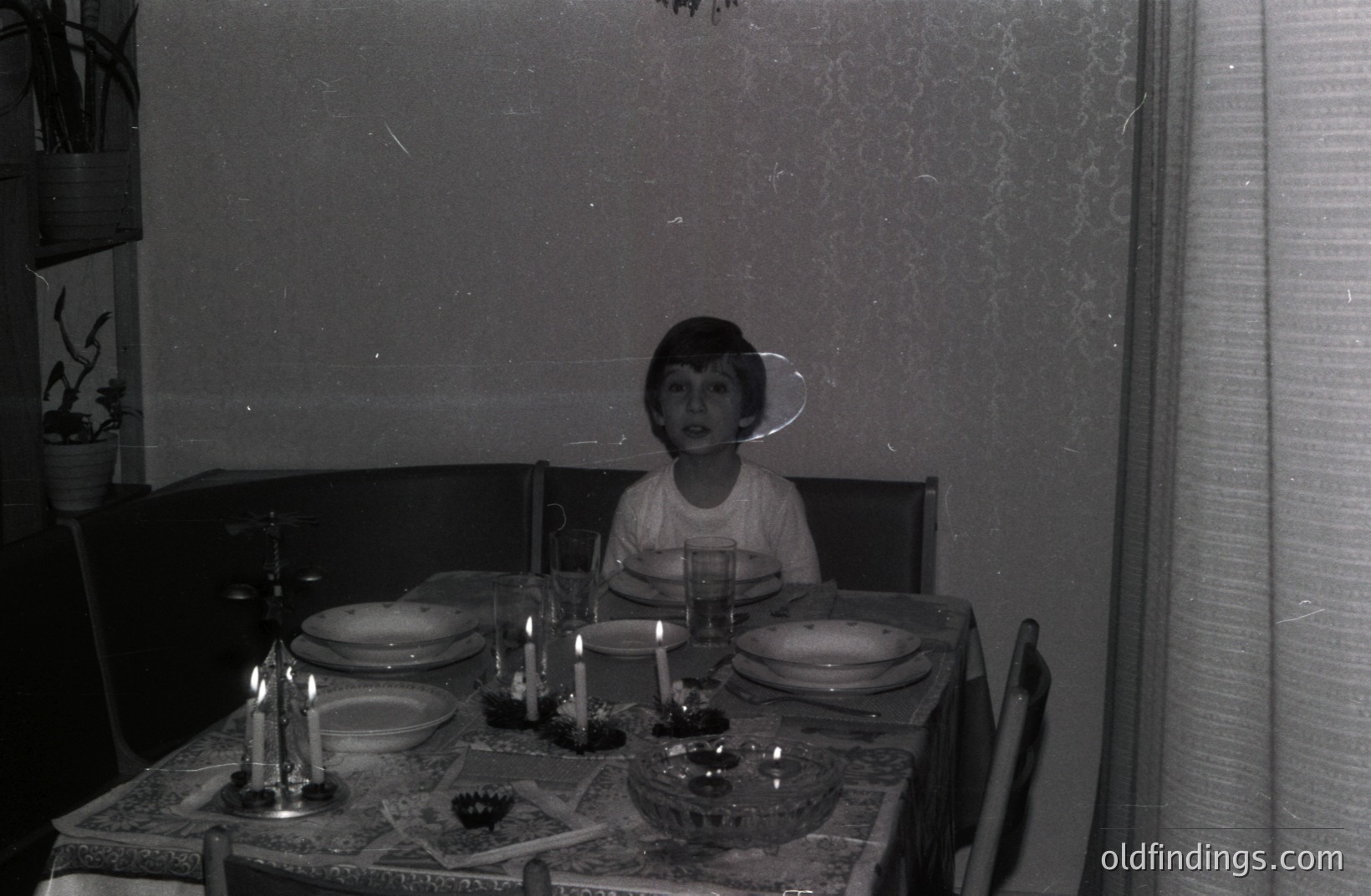 A young boy sits at a formally set table with multiple courses and lit candles. Textured wallpaper and dark furnishings suggest a domestic interior. The photograph's style and image quality are indicative of a 1960s or 1970s snapshot. Likely a family portrait.