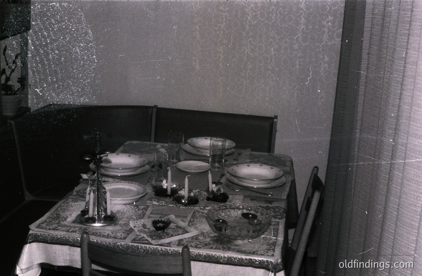 A formally set dining table occupies the scene, covered with a patterned tablecloth. Four place settings feature plates, glassware, and silverware. Candles and decorative accents enhance the ambiance. Likely a mid-century domestic interior, potentially . Photograph shows age and wear.