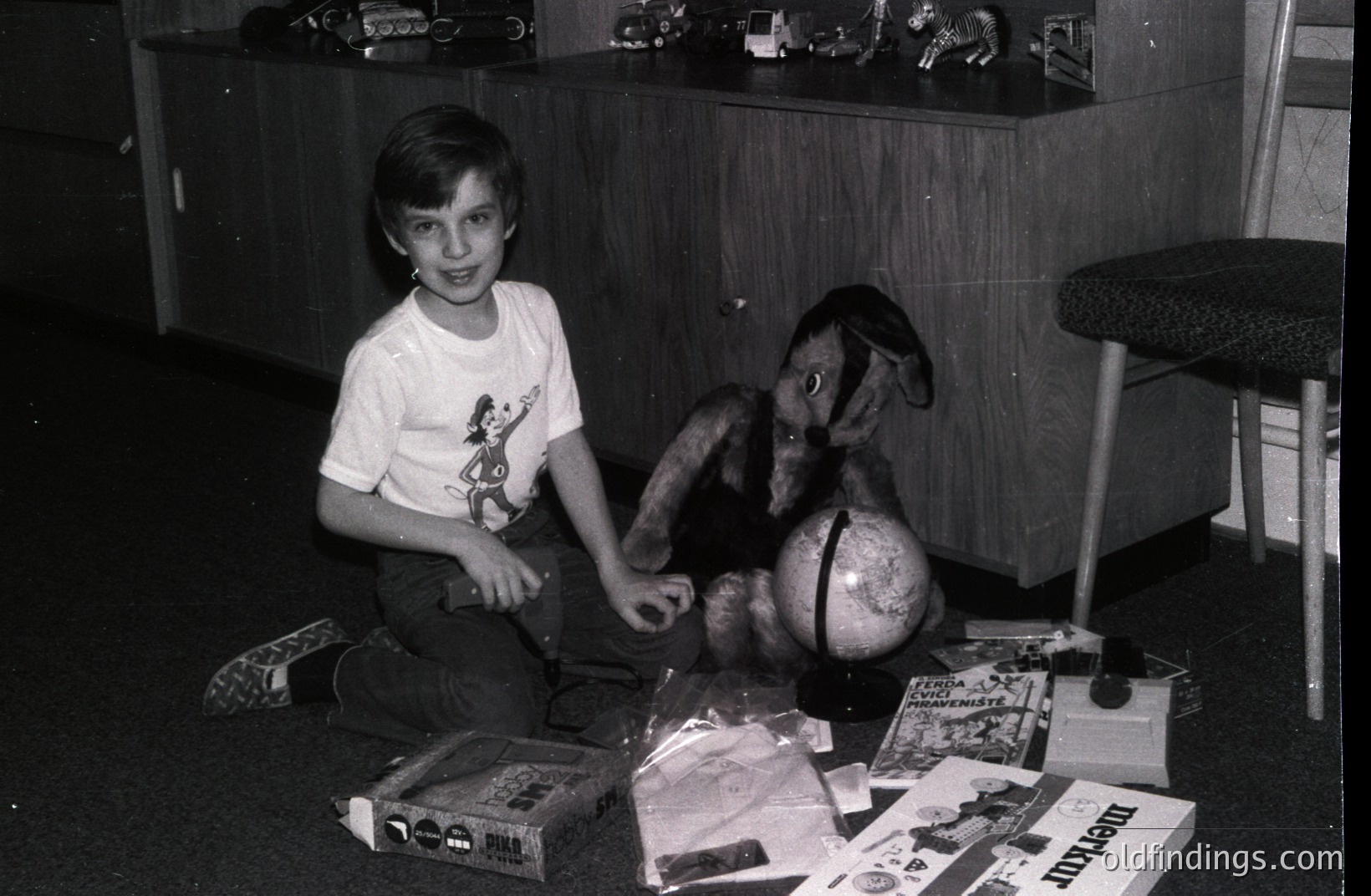 Young boy seated amid toys & gifts, including a plush deer and globe, surrounded by wrapped presents. Appears to be a domestic scene, likely a birthday or holiday celebration. The style suggests a 1970s timeframe. Simple, mid-century furniture adds to the period aesthetic.