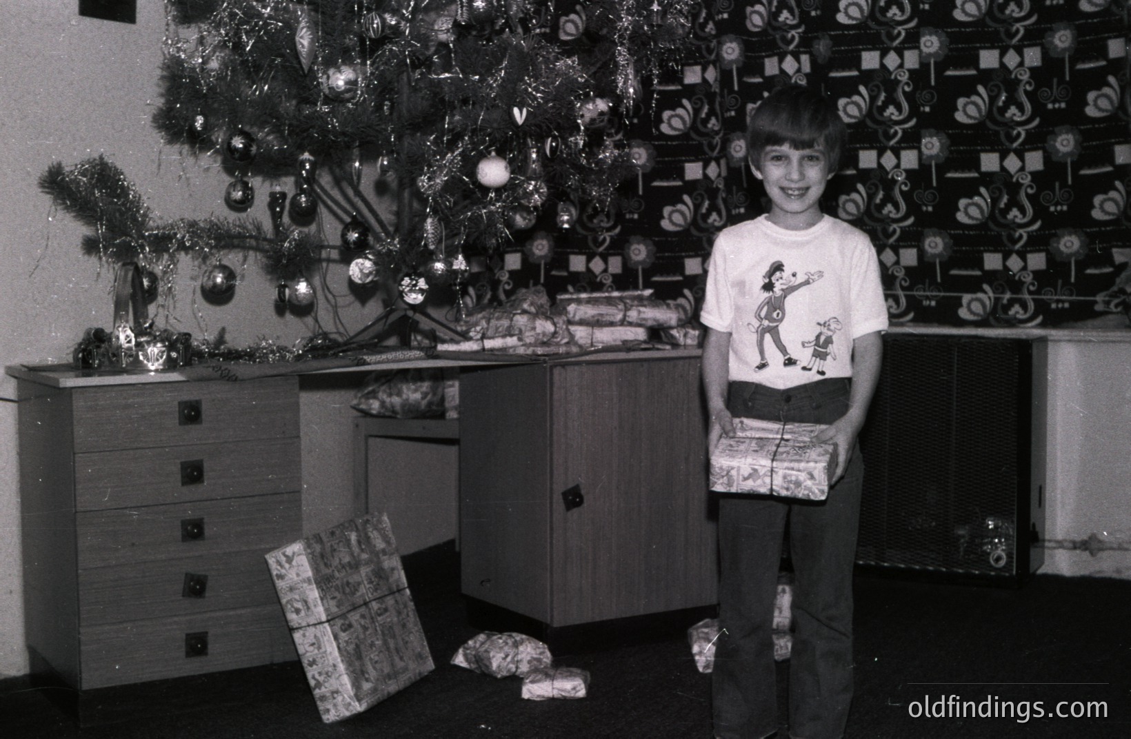 A young boy, likely in the 1970s, stands proudly holding wrapped gifts in a festive setting. A fully decorated Christmas tree with tinsel and ornaments fills the background. The patterned wallpaper & furniture suggest a mid-century home interior. A nostalgic, family-oriented portrait.