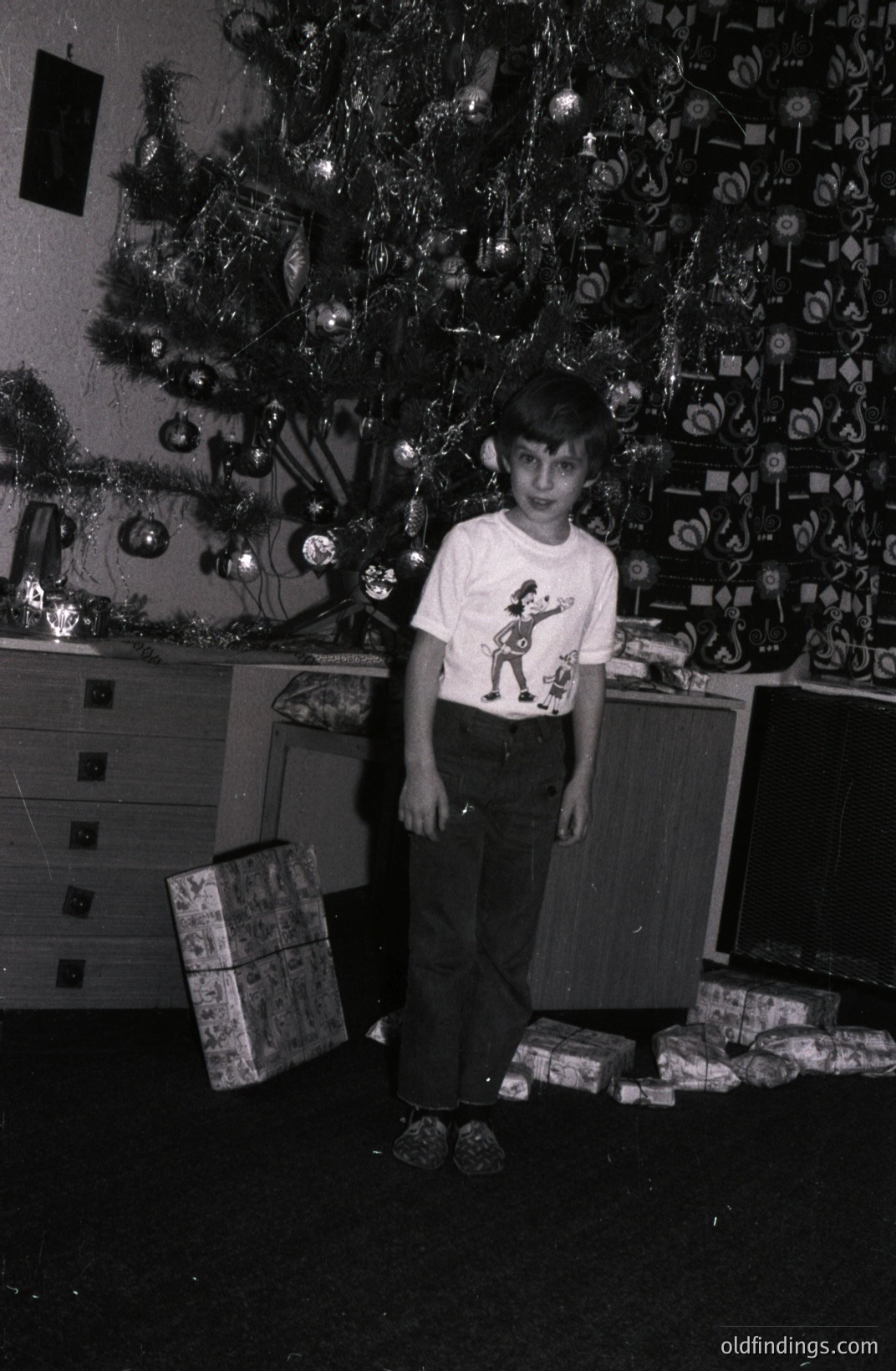 A young boy stands near a densely decorated Christmas tree, surrounded by wrapped presents. He wears a "Jetsons" themed t-shirt and bell-bottom jeans. Likely 1970s American scene, capturing mid-century holiday gifting traditions. A vintage piece of furniture sits behind him.