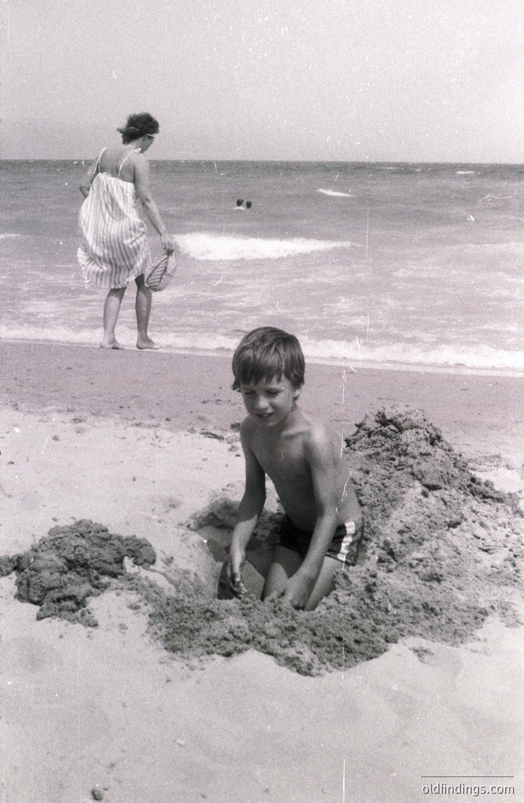 A young boy sits within a sand pit on a beach, looking toward the camera. A woman in a patterned dress stands further away, near the water's edge. The scene evokes a candid family moment, likely a vacation snapshot. Appears to be mid-20th century, possibly 1950s-1970s. Valuable for nostalgic/lifestyle imagery.