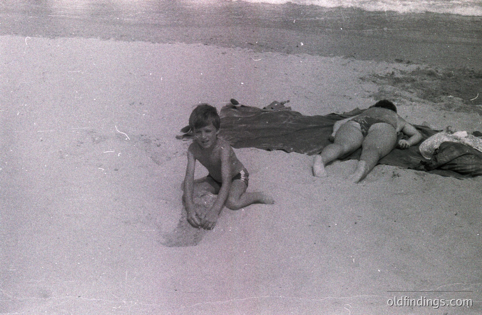 Two boys in swim trunks relax on a sandy beach, one playing with sand, the other resting on a blanket. Grainy black and white image suggests a candid, informal moment, possibly a family vacation. Likely 1960s-70s. Potential use for design or nostalgia-themed stock.