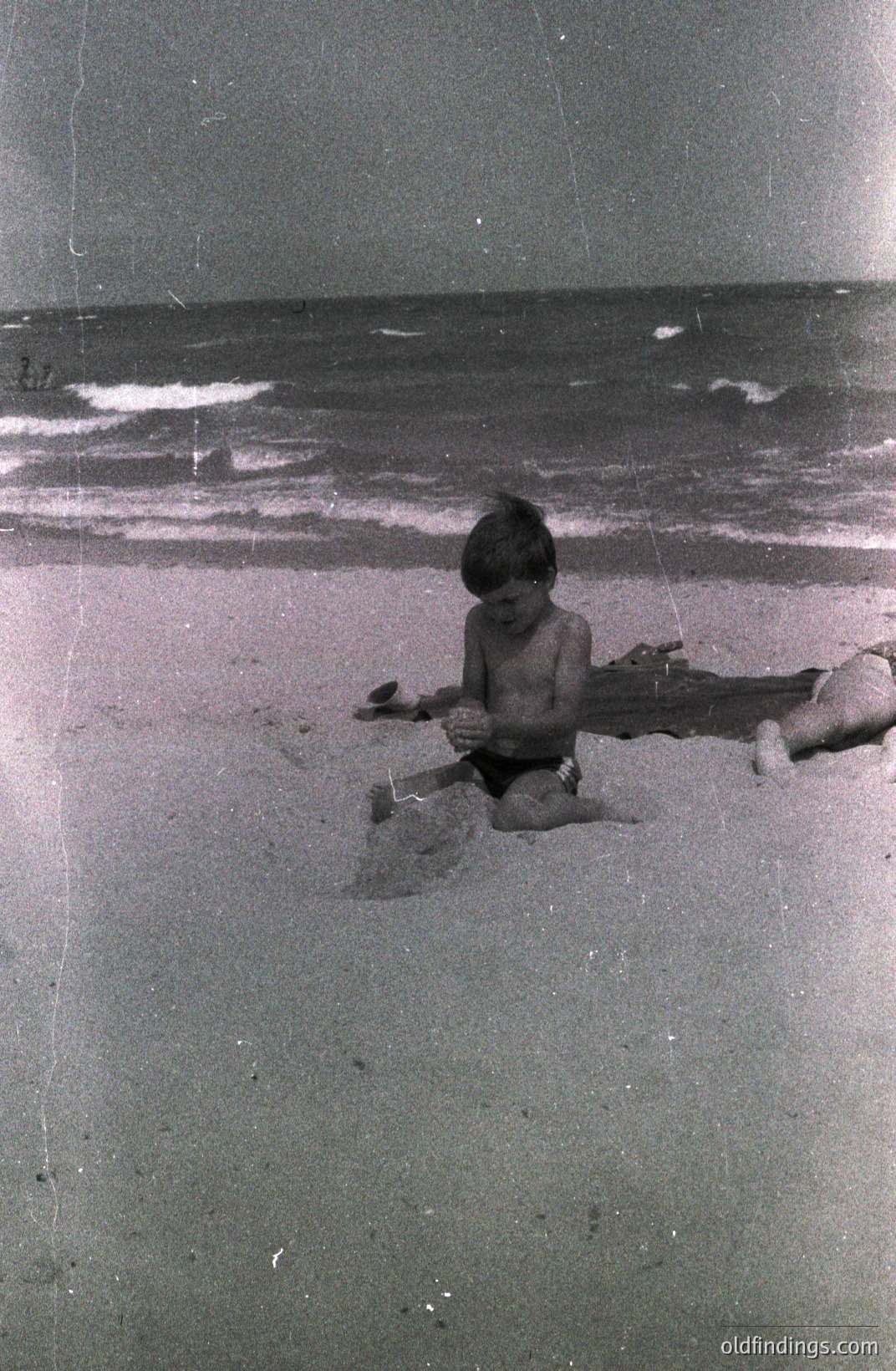 A young boy, lightly tanned and shirtless, kneels on a sandy beach, using a plastic shovel to build something. A log lies partially submerged nearby. Gentle waves meet the shoreline, extending to a gray sky. Likely 1960s-1970s era, possibly a family vacation snapshot.