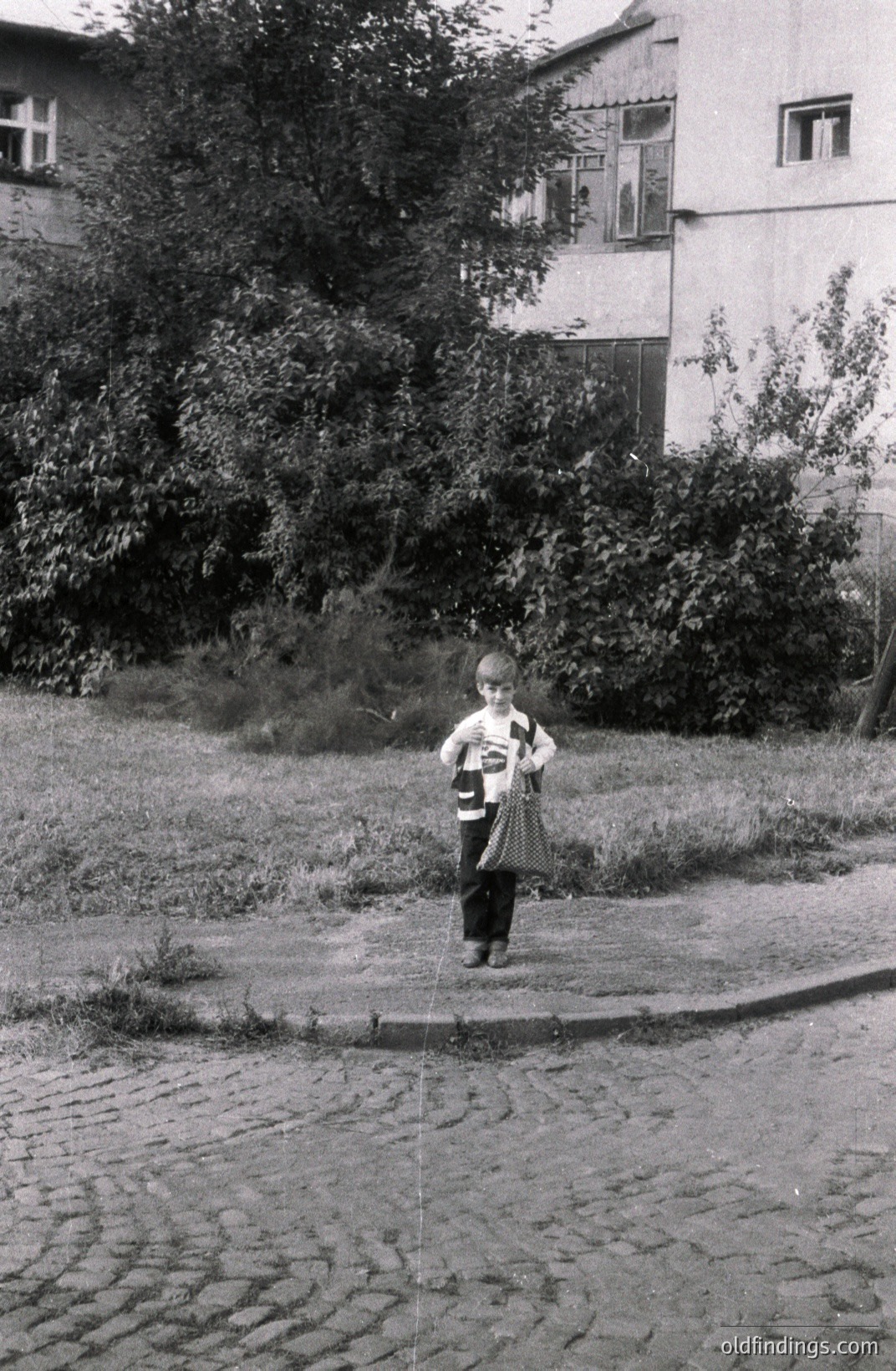 A young boy stands on a cobbled street, framed by dense foliage and a modest, multi-story building with distinctive window frames. He wears a patterned shirt and dark trousers, holding a small bag. Likely Eastern Europe, mid-20th century, showcasing vernacular architecture and everyday life.