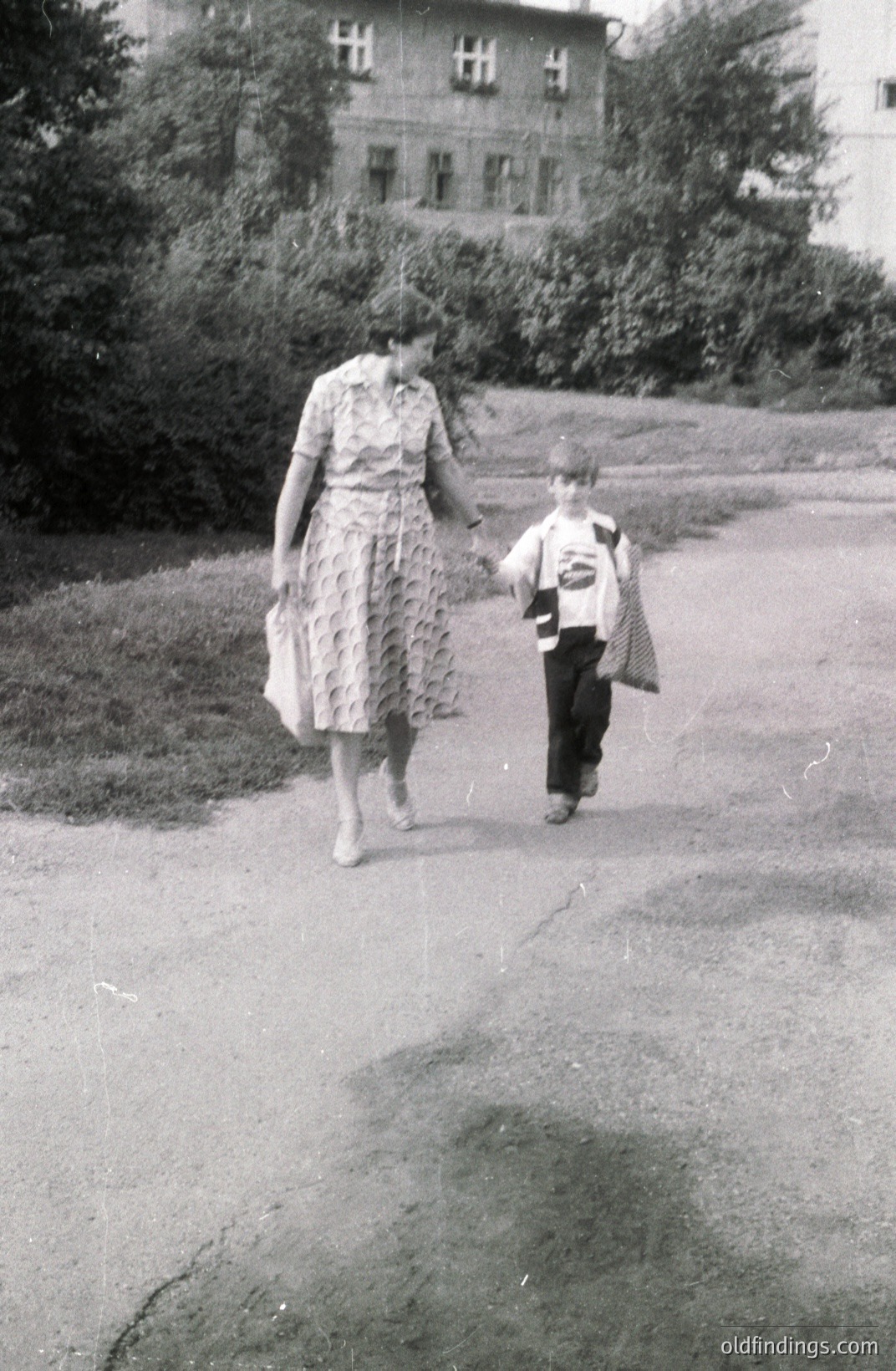 A woman and young boy walk hand-in-hand along a paved path. She wears a floral print dress; he carries a satchel. A large, multi-windowed building is visible in the background. Likely a snapshot from the 1960s or 70s. Evokes mid-century family life.