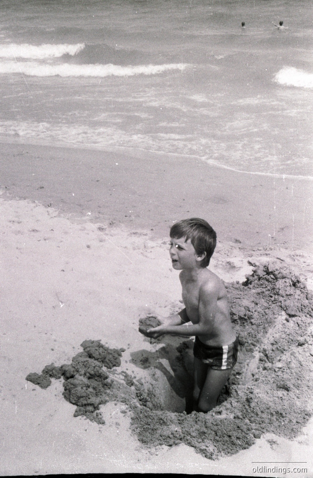 A young boy, possibly 7-10 years old, sits within a shallow pit he's dug on a sandy beach. He wears patterned swim trunks, holding a handful of wet sand. The backdrop shows a gentle, wave-swept coastline. Likely a candid snapshot from the 1960s or 70s.