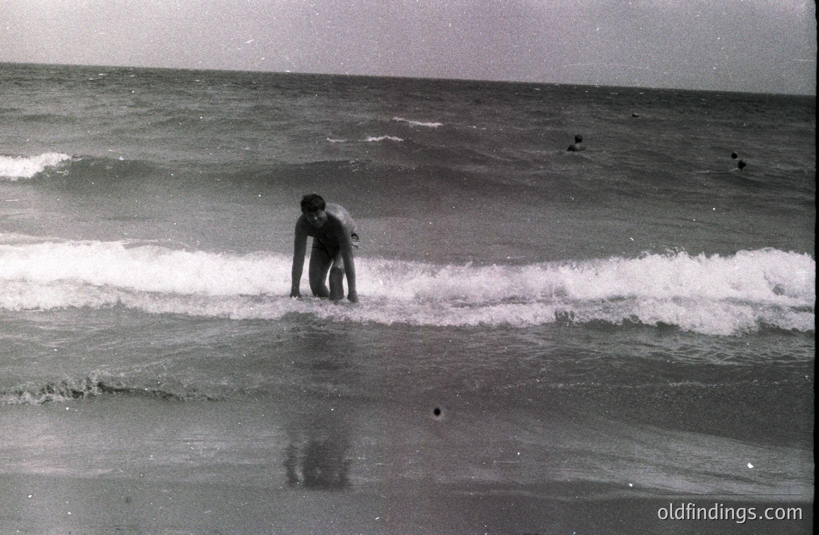 A lone figure wades through shallow, turbulent waves, appearing small against the vast seascape. Several distant figures are visible further out. The grainy, black and white image suggests a candid, possibly vacation snapshot. Likely mid-20th century.