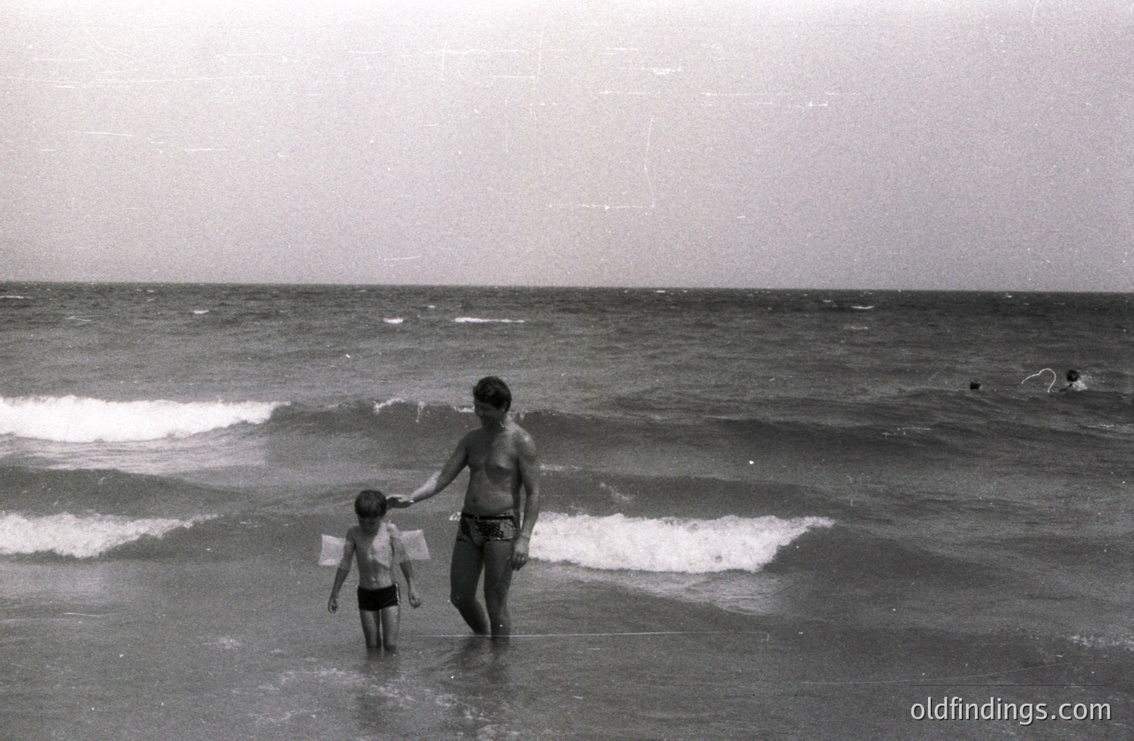 A man and young boy wade through shallow water along a sandy beach. The man holds the boy's hand. Waves gently break in the background; sparse figures are visible further out. Likely a family vacation snapshot. Photo’s quality suggests a 1970s origin. Evokes nostalgia and a sense of carefree summer days.