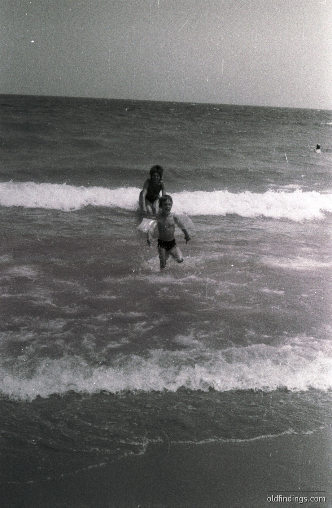 A boy wades into breaking waves, holding a large, inflatable beach toy resembling a swan. The image shows a mid-century beach scene with grainy, black-and-white film. Likely captured in the 1960s or 70s. Suggests leisure, childhood summers, and coastal vacations. Potential for stock photography/vintage design use.