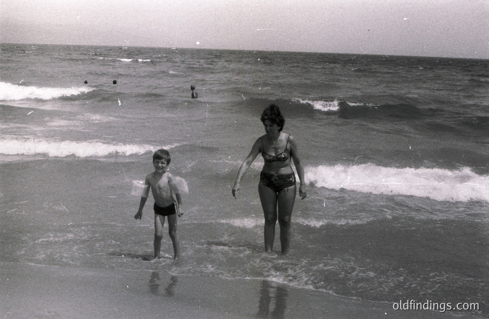 A mother and young son stand in shallow water along a sandy beach, waves gently lapping around them. The boy wears swim trunks, the woman a dark-colored bikini. Several figures are visible further out in the ocean. Likely a family vacation snapshot, circa 1960s-1970s.