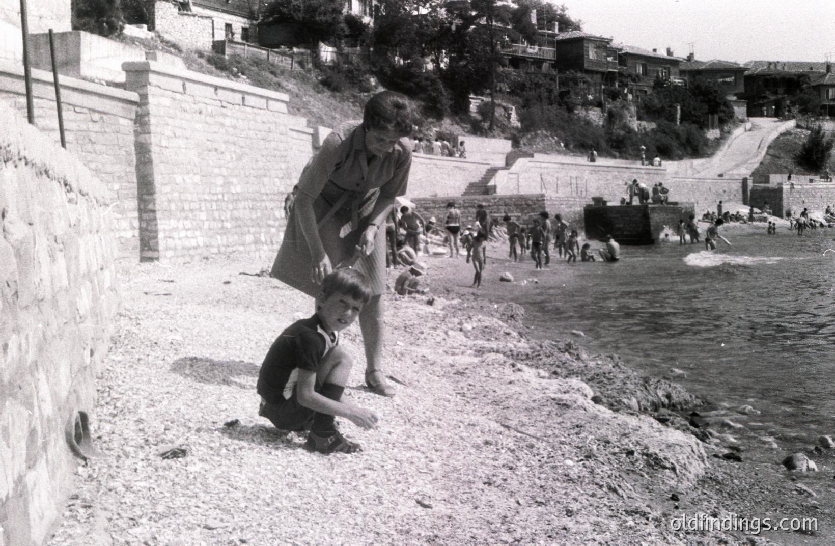 A young boy kneels on a pebbled beach, inspecting the ground, while a woman in a patterned dress watches nearby. A group of bathers are visible further out in the water and on steps leading up a stone embankment. Likely a seaside resort, possibly in Eastern Europe.