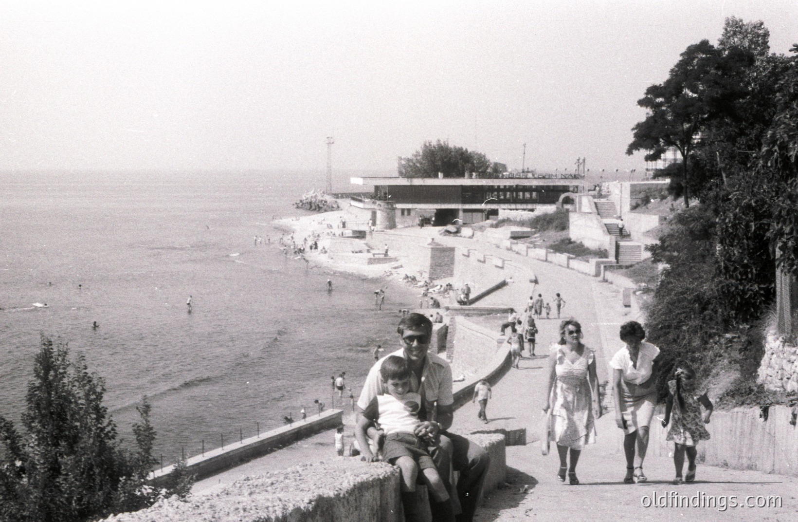 A black and white view of the Varna seaside resort in Bulgaria. A man cradles a child on a concrete retaining wall overlooking a sandy beach and ocean. 1970s architecture, a large, modernist concrete building dominates the background. Several people are strolling along a paved path. A coastal scene showing typical vacation leisure.