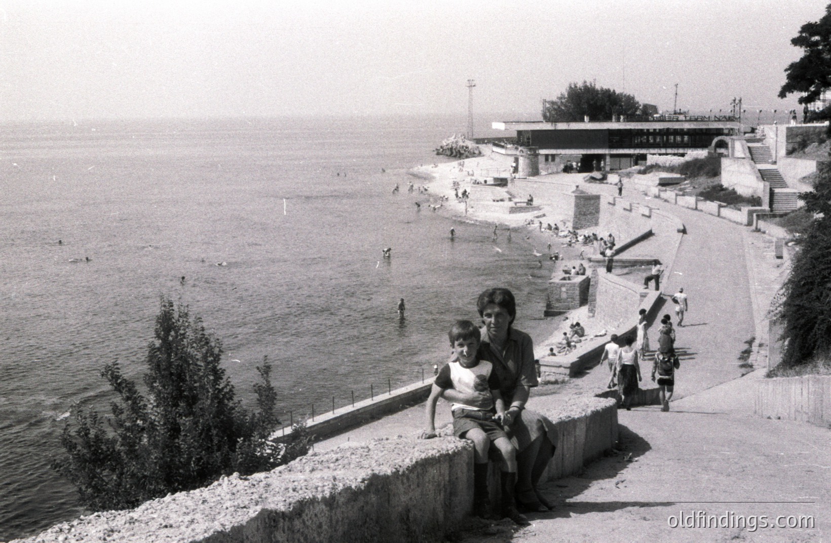 A family scene by the seaside: a mother and son sit on a stone retaining wall overlooking a sandy beach and the Black Sea. The architecture suggests a 1970s resort, likely in Bulgaria, with a prominent concrete structure with a curved roof. Numerous bathers enjoy the water.