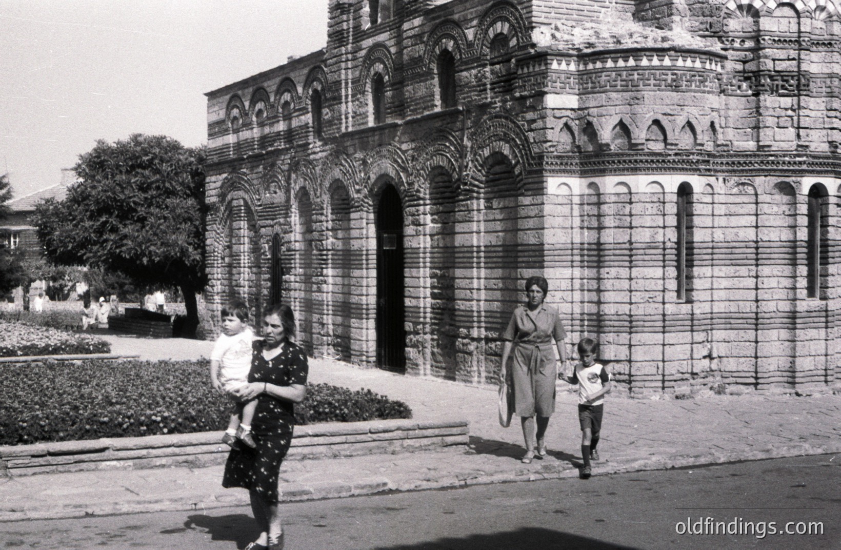 Striking b&w image: A family—mother carrying a child, another child running—pass before an ornate stone building with layered arches and a distinctive roofline. Likely Eastern European, possibly Bulgaria. Architecture suggests 1960s or 70s. Fine for architectural reference & nostalgia.