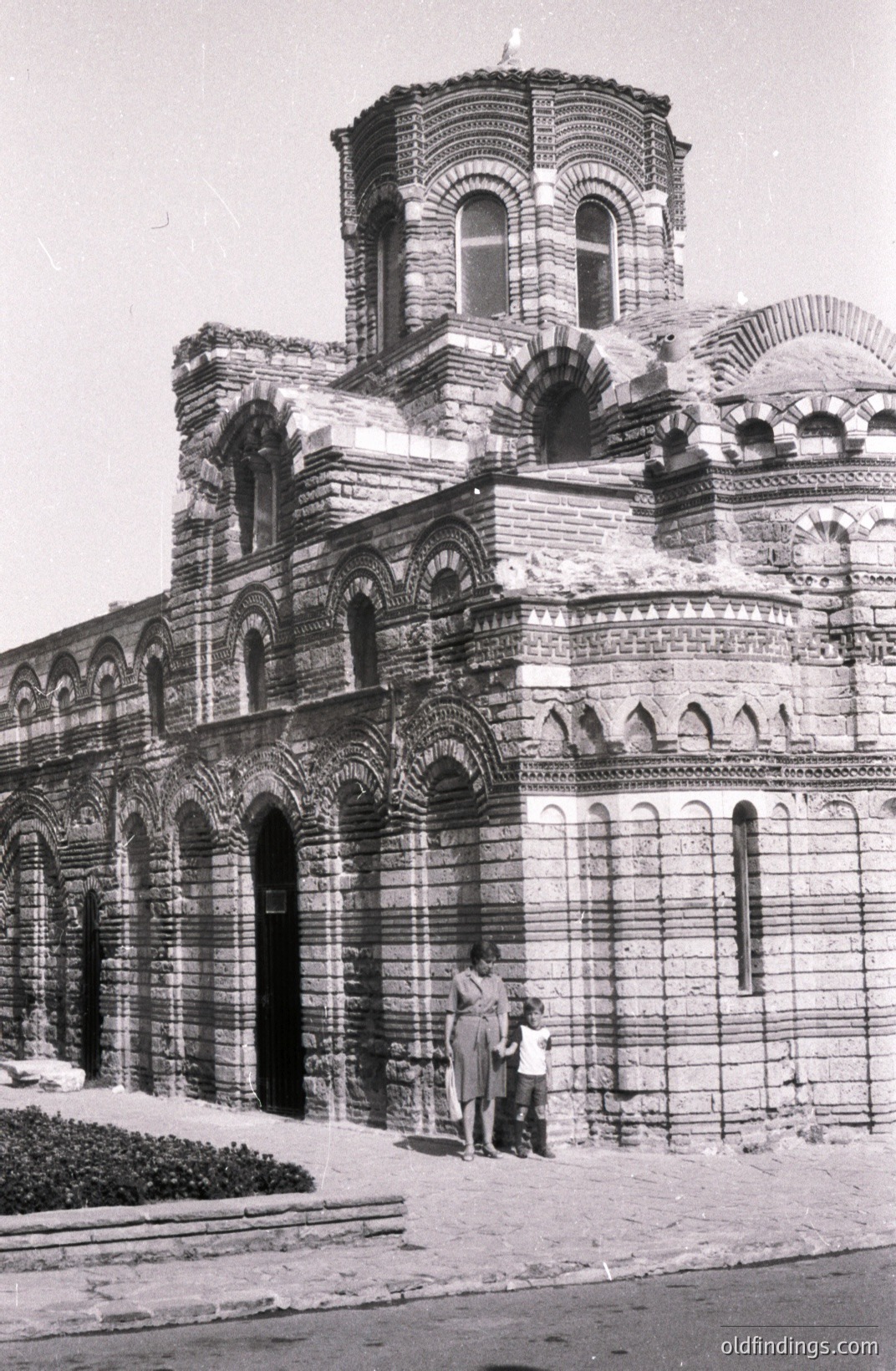 Striking architectural detail of the Aladağ Church, near Varna, Bulgaria. This 14th-century stone structure showcases distinctive arches and brickwork. Two figures, likely tourists, stand before the facade. Likely a 1960s travel photo.