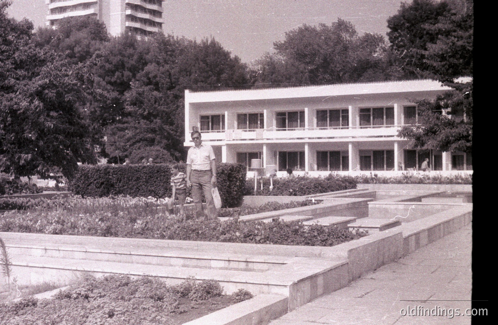 A man in trousers and a short-sleeved shirt stands near tiered landscaping featuring low shrubs and stone edging. Modernist architecture with balconies is visible beyond the greenery. Likely a resort or government building, possibly 1960s-1970s. Black and white film aesthetic.