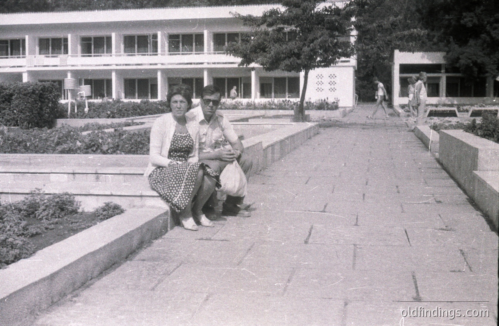Black and white photo depicts a couple seated on a low wall bordering a formal garden path. Behind them, a modernist seaside resort building with multiple balconies is visible. Likely Bulgaria, 1970s. Demonstrates post-war tourism architecture and fashion. A classic vacation scene.