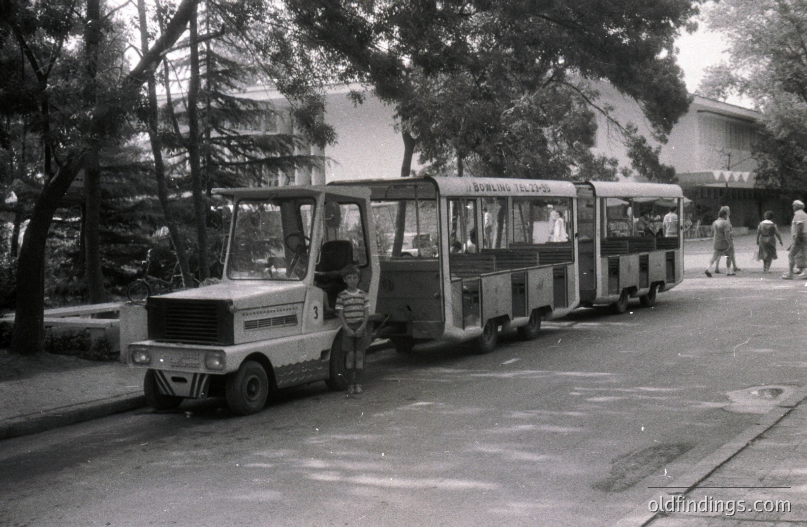 A vintage, open-air trolley or miniature train car stands on a paved road, a young boy stands near the front. The vehicle is labeled “BOWLING TEL-2M-56.” A large, modernist building is visible in the background, suggesting a resort or recreational complex. Likely Bulgaria, 1960s-1970s.