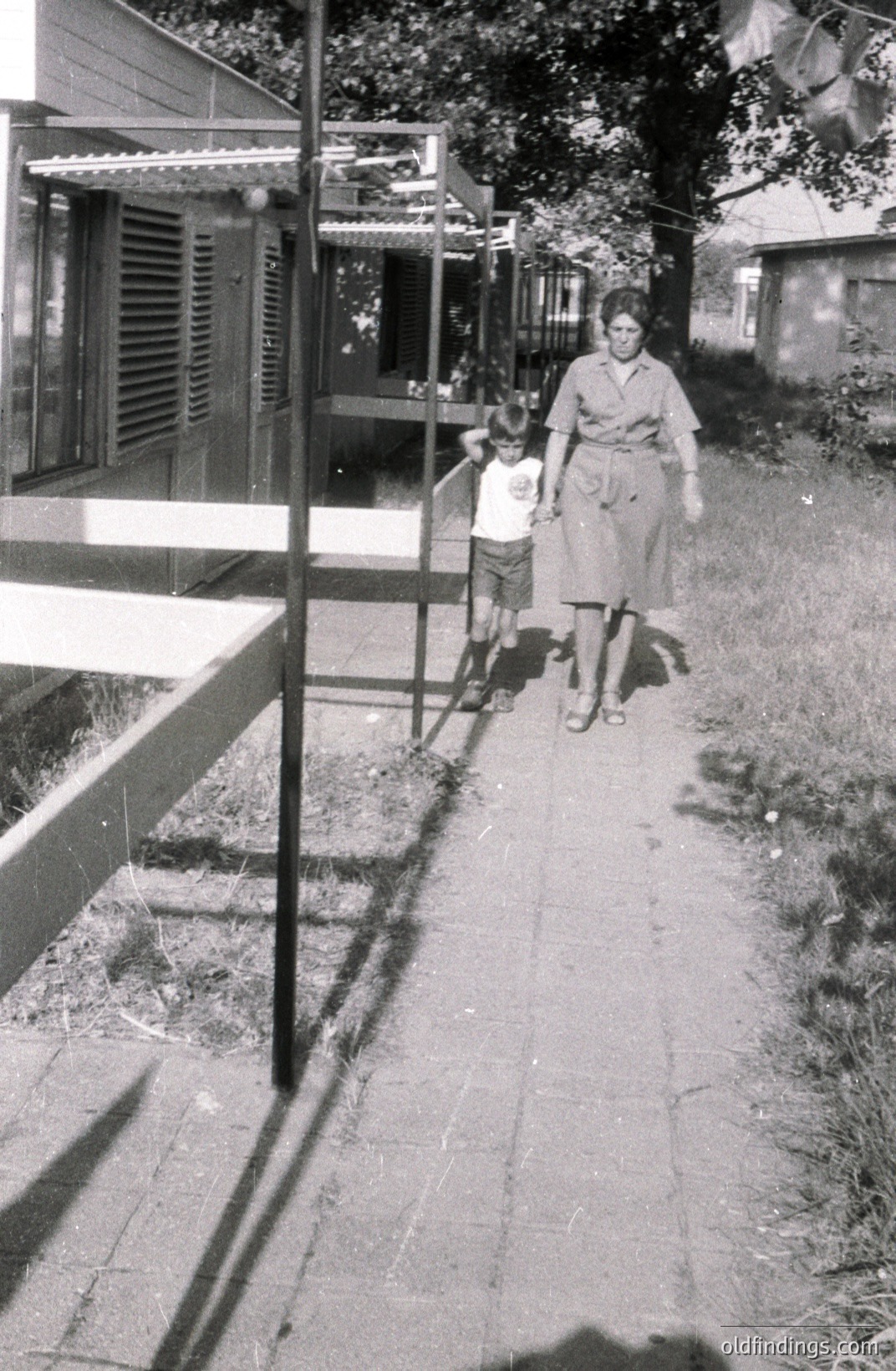 Black and white photo depicting a woman in a mid-length dress and a young boy walking along a brick-paved path. A building with shutters is visible in the background. Likely captured in the 1960s-70s, evoking a nostalgic domestic scene. Potential stock photo reference for retro lifestyle imagery.