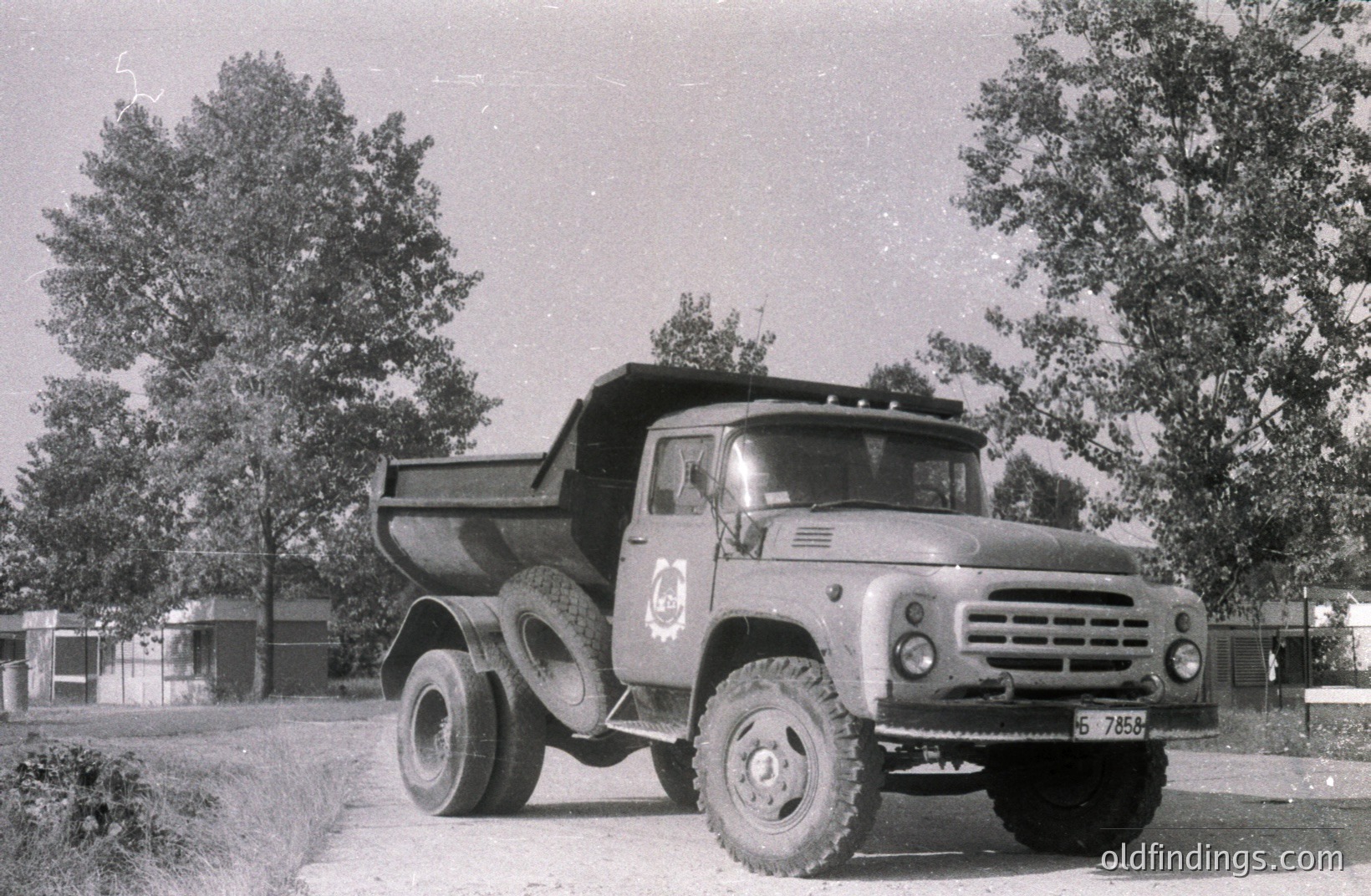 Heavy-duty Soviet-era dump truck, likely a Ural model, parked on a gravel road amidst sparse vegetation. Features include a prominent cab, robust tires, and a visible license plate. Likely a work vehicle in an industrial or agricultural setting. Circa 1960s-1970s Eastern Europe.