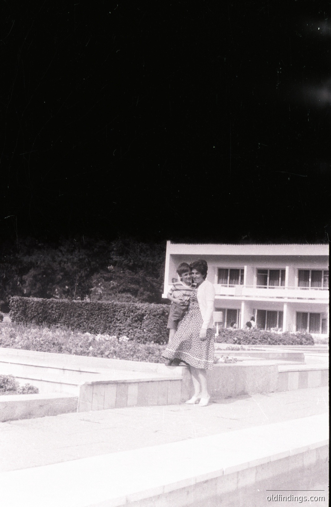 A woman carries a child outdoors in front of a modernist building, possibly a hotel or apartment complex. Features include a tiered facade & large windows. Likely 1960s-70s period, style aligns with mid-century architecture. Possible seaside resort location. Image displays a sense of quiet leisure.
