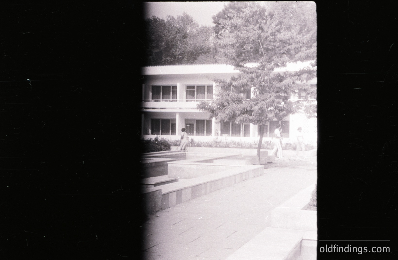 A monochrome photo showing a modernist building with large windows, viewed from a paved courtyard. Two figures are present, one seated near steps. Architectural style suggests mid-20th century, possibly Eastern European. Strong vignette effect due to film edge. Potential stock usage for design references or historical context.