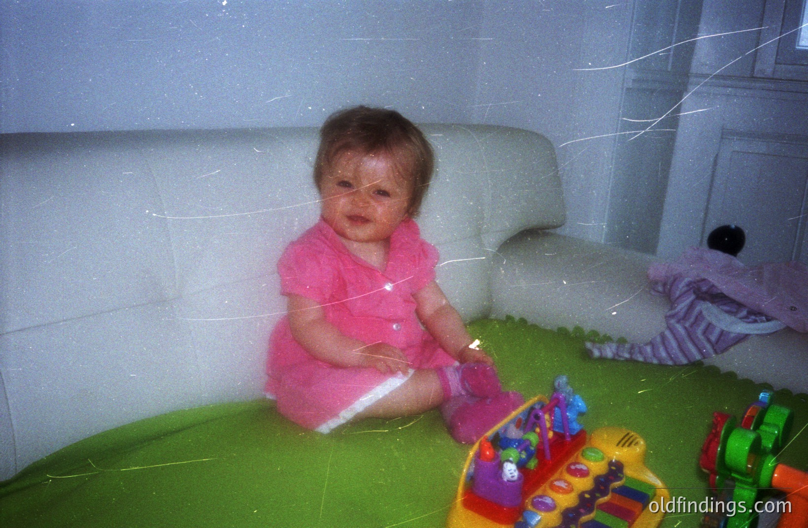 Infant sits on a green play mat, wearing a pink dress, surrounded by colorful toys. Appears to be a candid, indoor shot, likely from the 1990s or early 2000s, judging by the film aesthetic & toy design. The image exhibits visible film grain & light leaks.