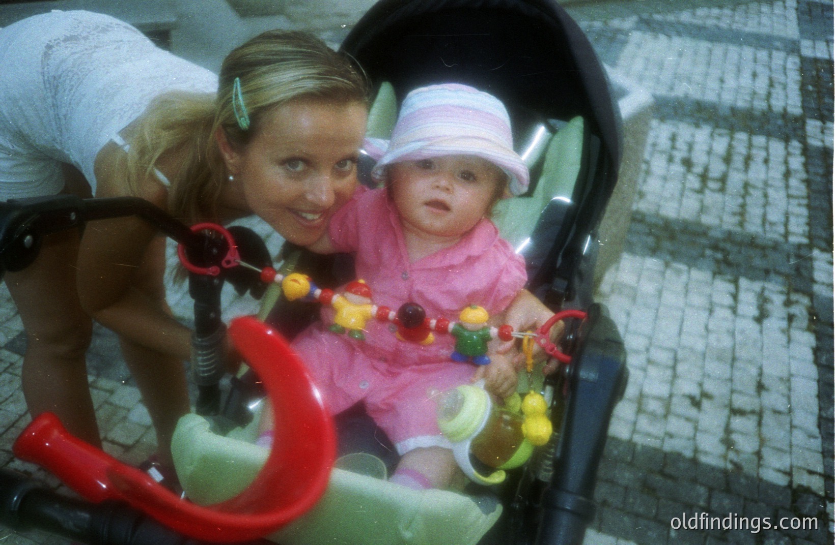 A young woman with blonde hair, styled in a high ponytail, gazes down at a baby seated in a vibrant green stroller. The baby wears a pink hat and pink dress, gripping a colorful toy. Appears to be a candid moment, likely a family outing. Evokes a 1980s/1990s aesthetic.
