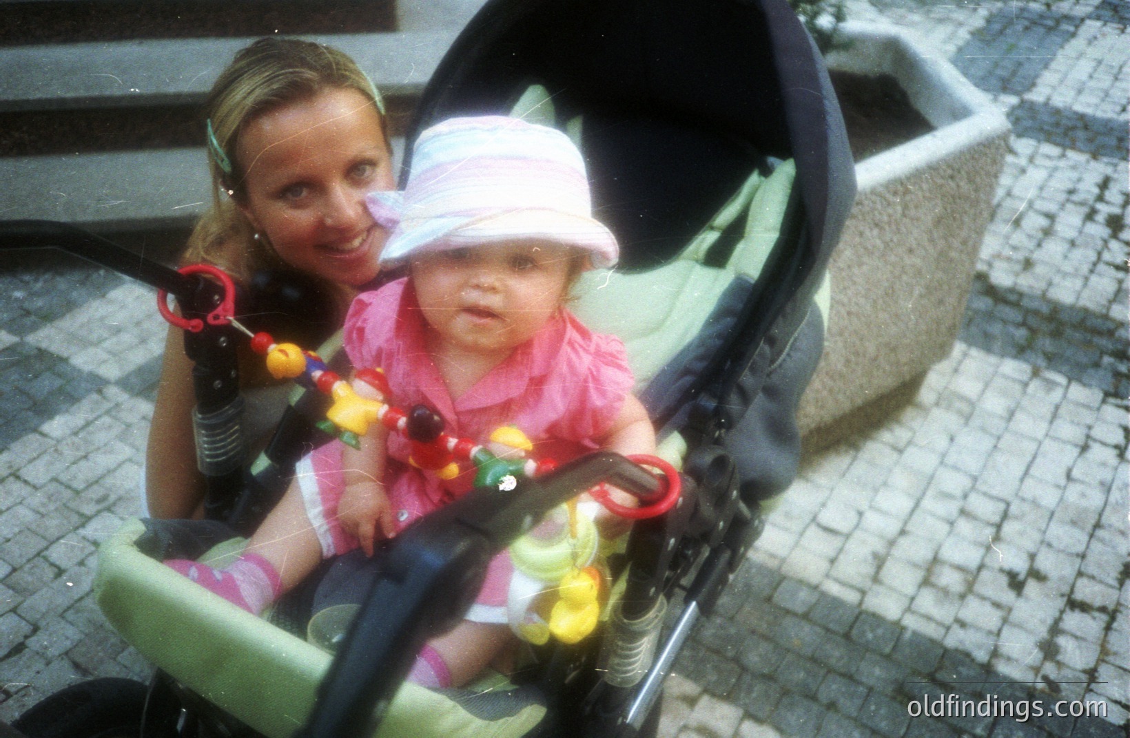 A baby in a stroller, wearing a pink hat and dress, gazes forward, held by a woman smiling in the background. A toy hangs from the stroller handle. The image appears to be from the 1990s, based on the style and color palette. Likely captured outdoors on a paved surface.