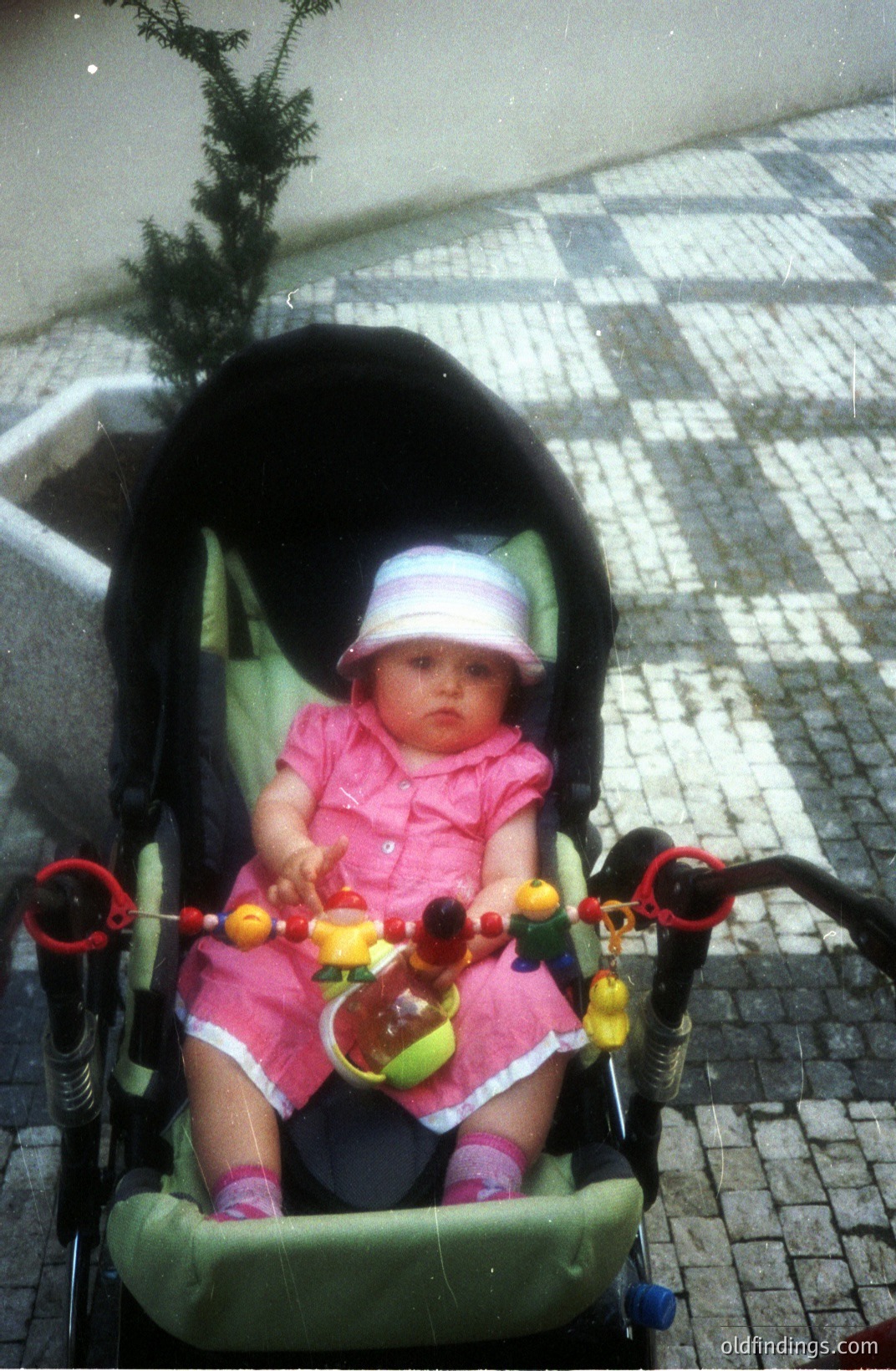 A baby, wearing a pink dress and a matching hat, sits in a green stroller with a mobile of toys attached to the handles. Seated outdoors on a paved area. Likely 1970s or 80s, judging by the stroller's style. Charming, nostalgic family portrait.
