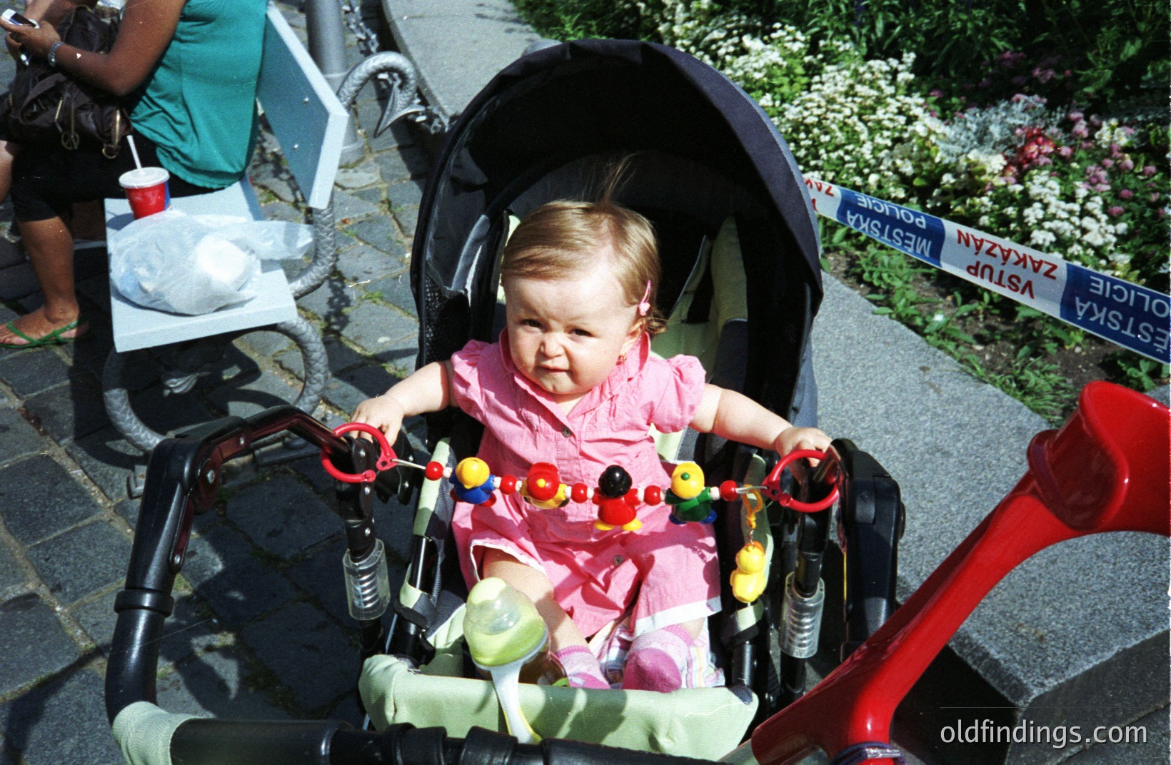 Infant seated in a stroller, gazing forward. Pink outfit and a playful, colorful toy hanging from the stroller's frame. Sign in Polish indicates a restricted area. Likely a candid moment, possibly 1990s or early 2000s.
