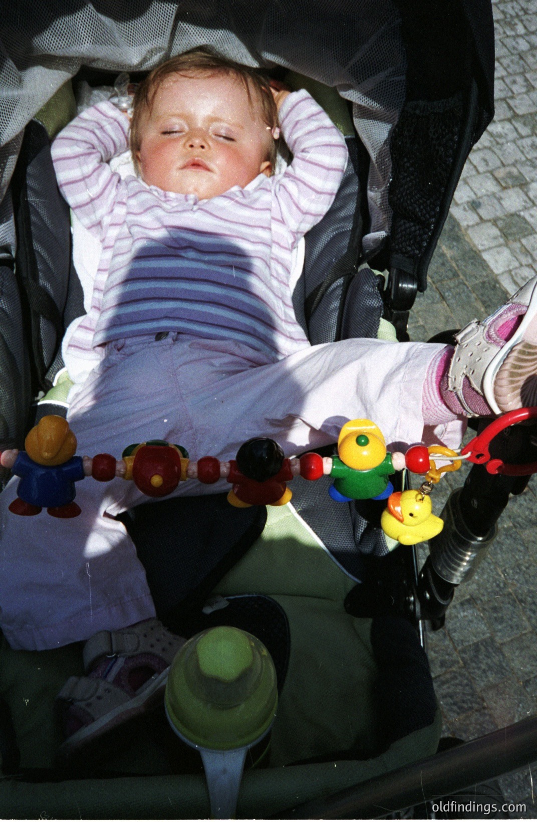Infant sleeping peacefully in a stroller. Wearing a striped onesie and socks. A colorful toy mobile with figures is attached to the stroller’s frame. Likely a candid moment captured outdoors, possibly 1990s or early 2000s. Could serve as stock photography for parenting, baby products, or lifestyle themes.