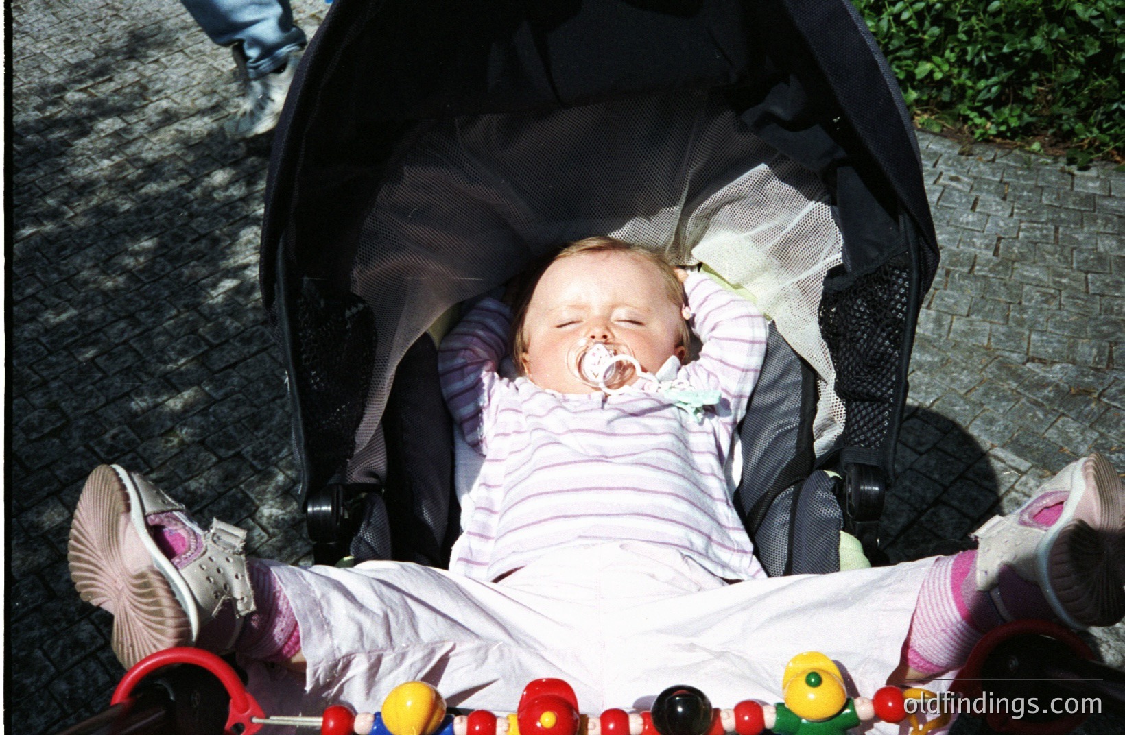 A baby lies asleep in a stroller, pacifier in mouth, wearing a pink and white striped outfit with socks and shoes. The stroller's netting is visible above. A toy rattle rests beneath. Appears to be a casual, candid moment.