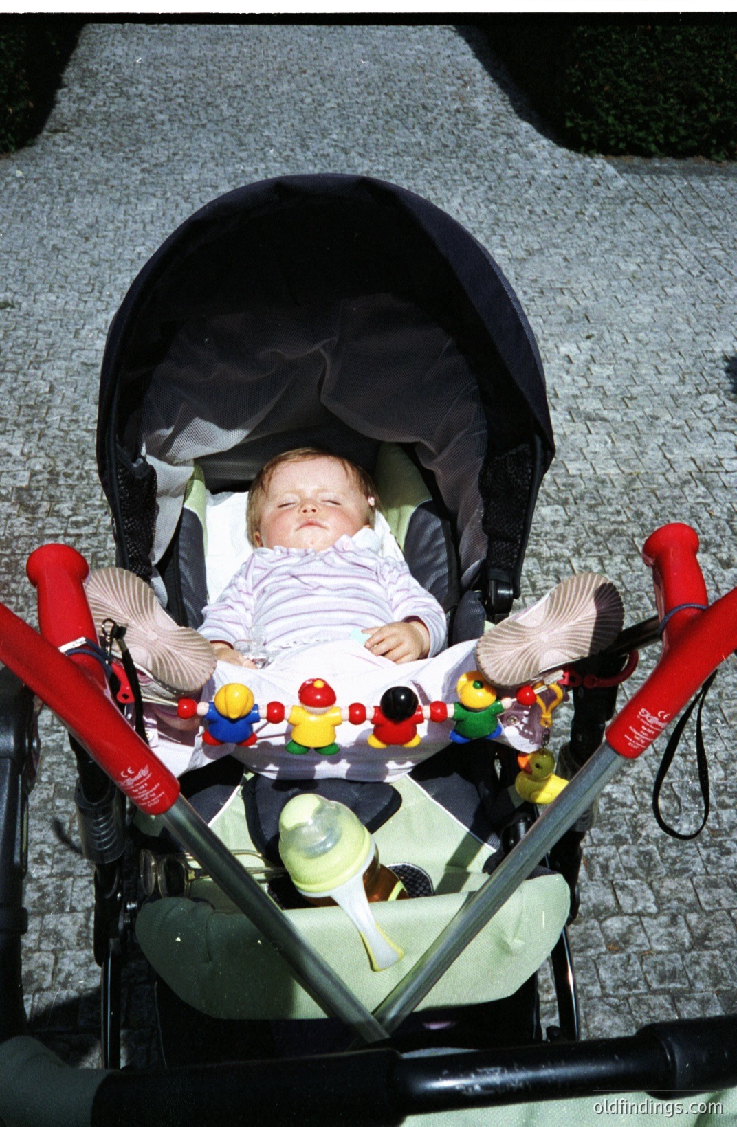 Infant asleep in a vintage stroller with a red-handled frame and patterned fabric canopy. A toy bar with dangling figures sits above. Appears to be a candid moment, possibly taken in the 1970s or 80s. Stock photography potential.